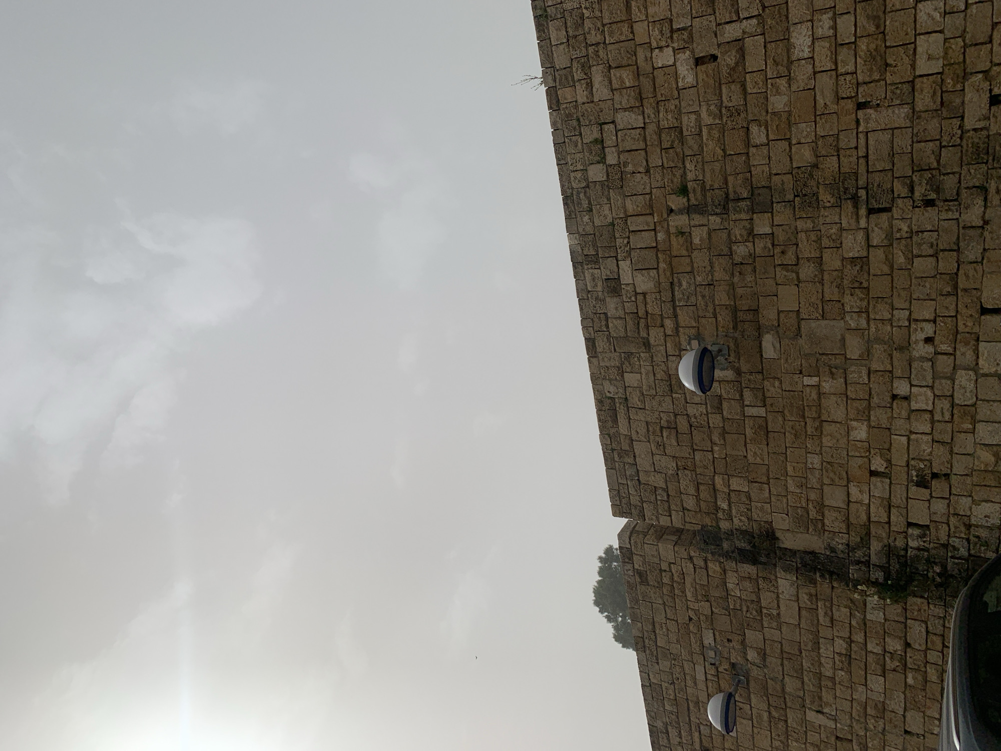 The image presents an upward perspective, dominated by a large, ancient-looking stone wall on the right, contrasting with an expansive, overcast sky on the left. The wall is constructed from varied rectangular and square-cut stones, exhibiting shades of brown, beige, and grey, with scattered patches of green moss or small plants growing within its crevices. Two modern, hemispherical outdoor lamps, white with dark blue or black bands, are attached to the stone structure.
Towards the middle-right, behind a section of the wall, the dark green foliage of a tree is visible. The sky is largely covered by diffuse, light grey clouds, with a brighter, almost white area in the bottom-left corner suggesting the presence of the sun, creating a lens flare effect. A small, dark, bird-like object is visible in the mid-left section of the sky. The very bottom-right corner shows a dark, reflective surface, likely a part of a vehicle such as a car.
The scene captures a blend of historical architecture and modern elements under a cloudy daylight sky. No specific activity or people are present. The impressive stone wall, combined with the given location context, suggests a historical part of Tel-Aviv, Israel, such as Old Jaffa.