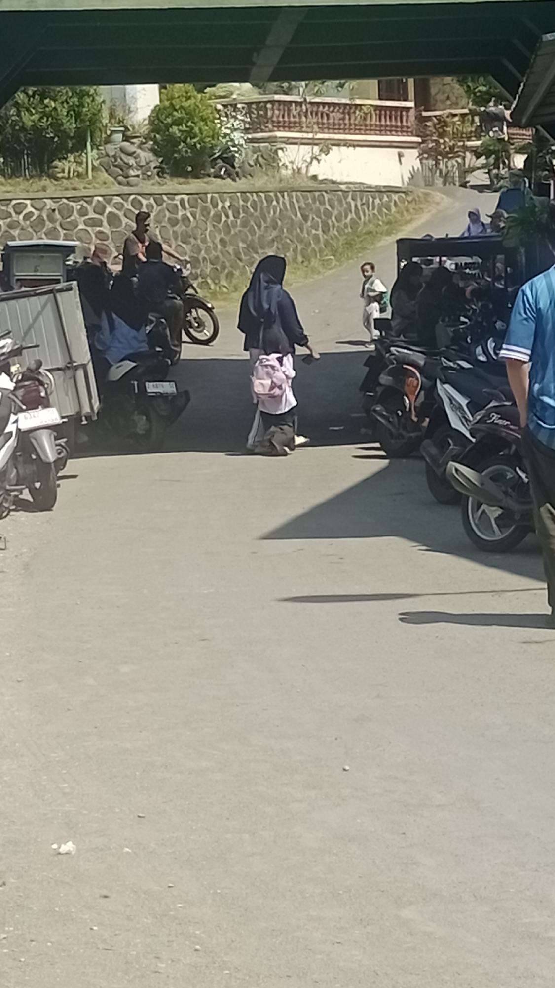 A group of people are gathered near parked motorcycles in an outdoor area. In the foreground, a person wearing a dark hijab and a pink backpack walks away from the camera. To the left, a white truck bed is visible with a person in a blue shirt standing nearby. Several motorcycles are parked in the middle ground, some with riders. In the background, a stone wall separates the paved area from a grassy slope with trees and a building. The scene appears to be a public space, possibly a parking lot or roadside area in Pakemitan, Indonesia, indicated by the style of motorcycles and the architecture in the background. The lighting suggests it is daytime.