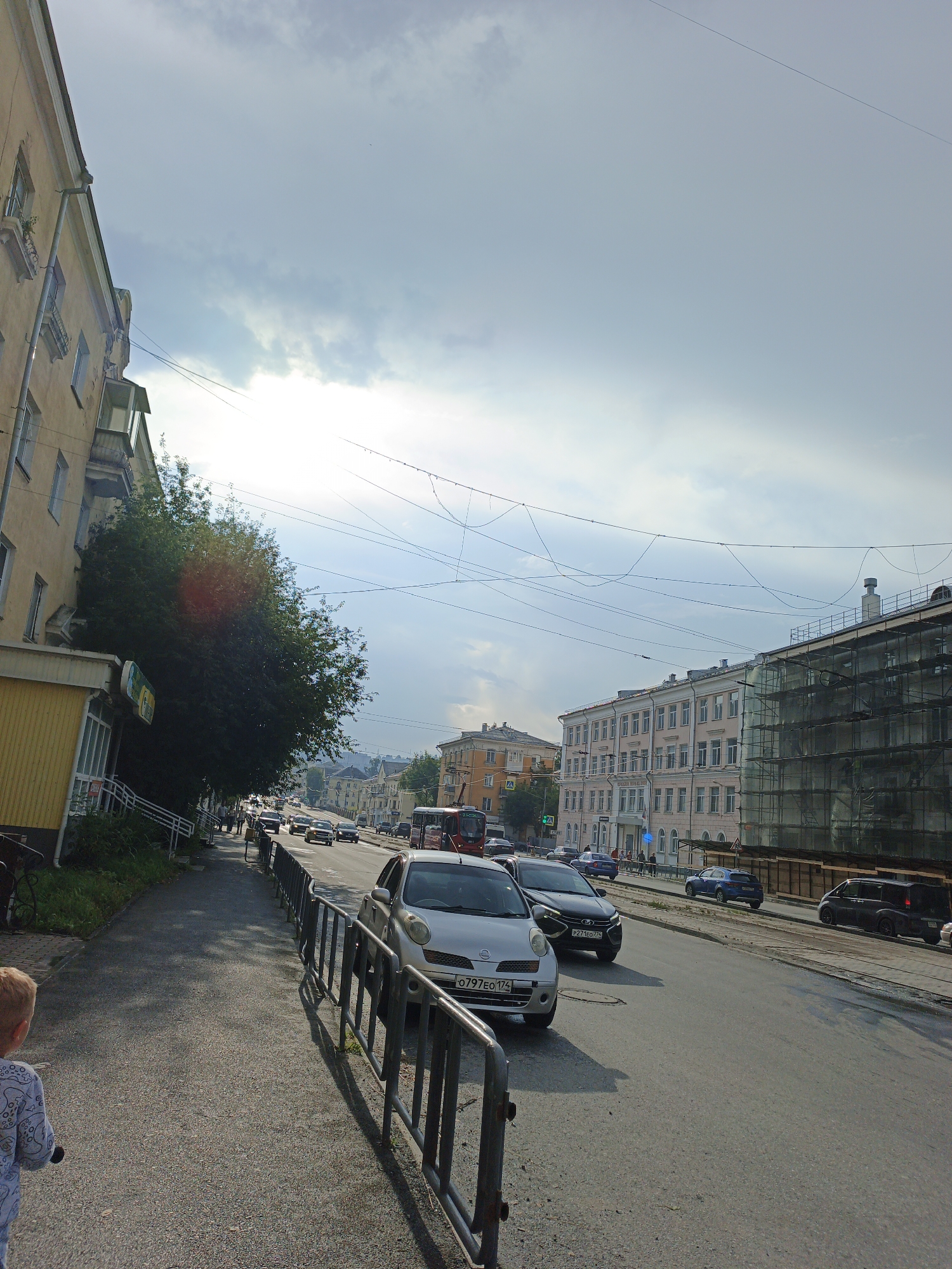 The image depicts a street scene in Zlatoust, Russia, under a partly cloudy sky. On the left, a pale yellow building with multiple windows and balconies runs along the sidewalk. A large, dark green tree is situated next to this building, casting shadows on the sidewalk. The sidewalk itself is paved with asphalt and has a black metal railing separating it from the street.
The street is a multi-lane road with several cars in motion, including a silver Nissan, a black sedan, and a blue SUV. Traffic appears to be moving at a moderate pace. Across the street, a pink building with many windows stands prominently, and next to it, a building is covered in scaffolding, suggesting construction or renovation. Further down the street, more buildings and a red trolleybus are visible.
A young child, seen from behind, stands on the sidewalk on the left side of the frame, facing the street. The overall impression is a typical urban environment on a day with mixed weather.