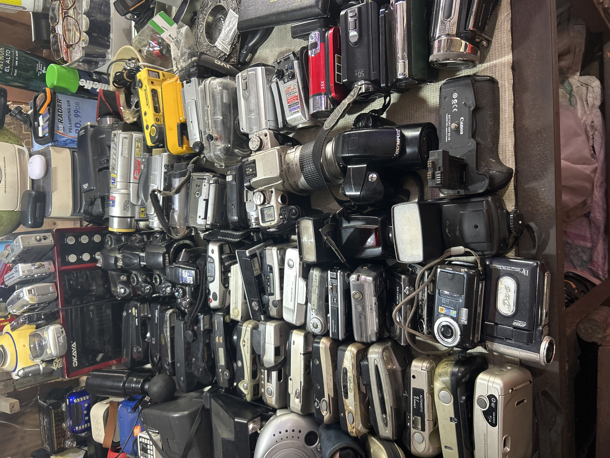The image shows a cluttered collection of old cameras and camcorders arranged on a table or shelf. The items are diverse, including digital cameras, film cameras, and video cameras of various brands and models. Some of the items appear to be in good condition, while others show signs of wear and tear. The background is dimly lit and indistinct, but it appears to be an indoor setting. The overall impression is one of a storage or collection area for electronic devices. There are no people visible in the image, and no specific actions or interactions are depicted. The image does not provide any specific visual cues that would indicate its location in Denpasar, Indonesia, or any other specific city. The items themselves are not particularly indicative of a specific geographical location.