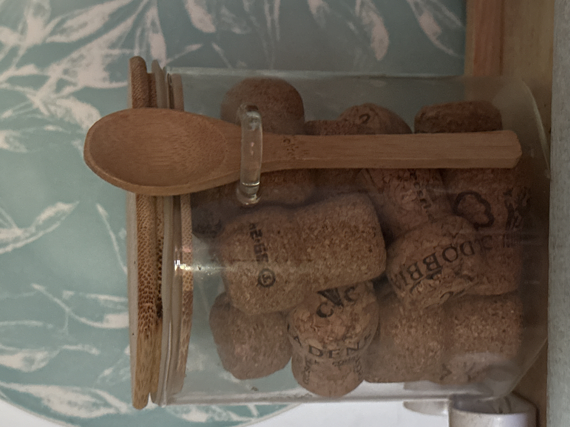 The image shows a clear plastic container filled with used wine corks. A small wooden spoon lies across the top of the container. The container is sitting on a white surface against a background of a blue and white patterned surface. The corks have various markings and text on them, with the brand "DOBBIA" visible on one of them. The exact location in the city of Zelenograd, Russia, is not identifiable from the image. There are no people in the frame.