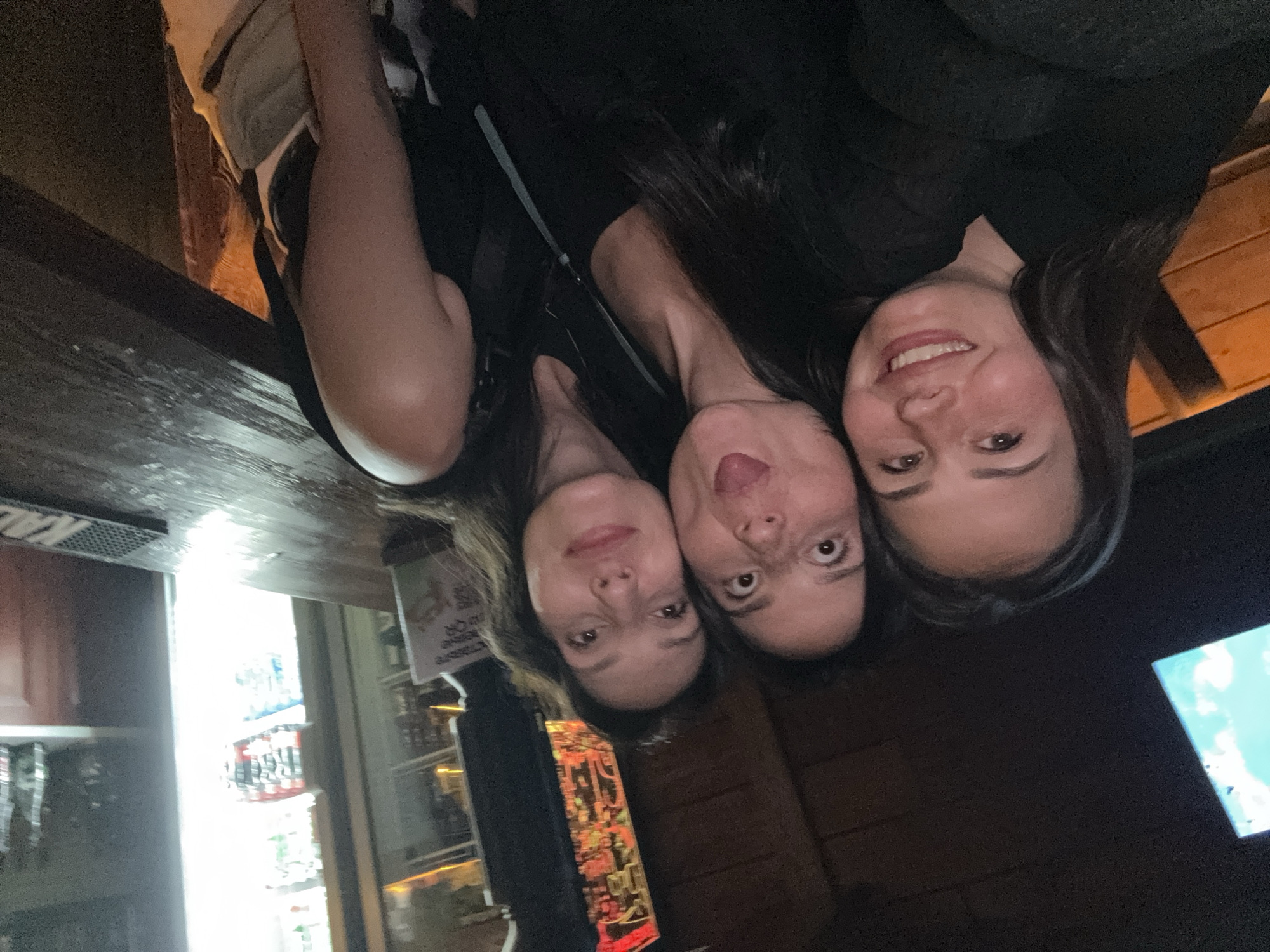 Three women are closely posed together, likely for a selfie. They are inside what appears to be a bar or restaurant, evidenced by the bar counter visible in the foreground, with a refrigerated display case behind it containing various beverages. The background is dimly lit with wooden walls. A television screen is partially visible in the bottom right corner.
Based on the prompt, this scene is located in the city of Chaikovsky, Russia.