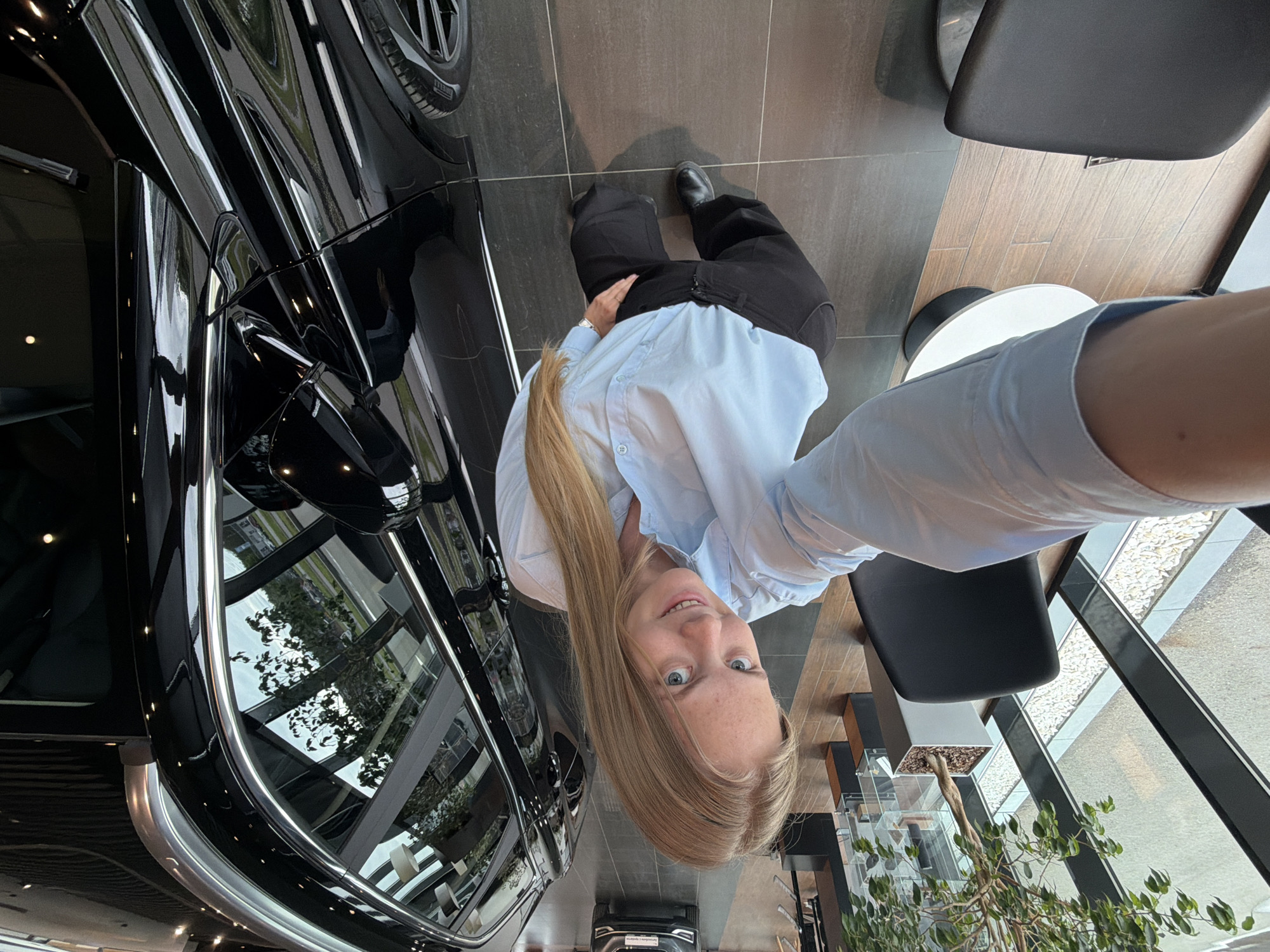 The image is a selfie taken from a high-angle perspective. A young woman with long blonde hair and blue eyes is the primary subject. She is wearing a light blue button-down shirt and dark trousers with black shoes. She is positioned near a shiny, black car with reflective windows that reveal the outside environment.
The location appears to be an indoor showroom with tiled floors. Several chairs and tables are visible, along with potted plants. The reflection in the car's windows suggests an external view of modern buildings and greenery. The image was not taken in the city of Izhevsk, Russia.
