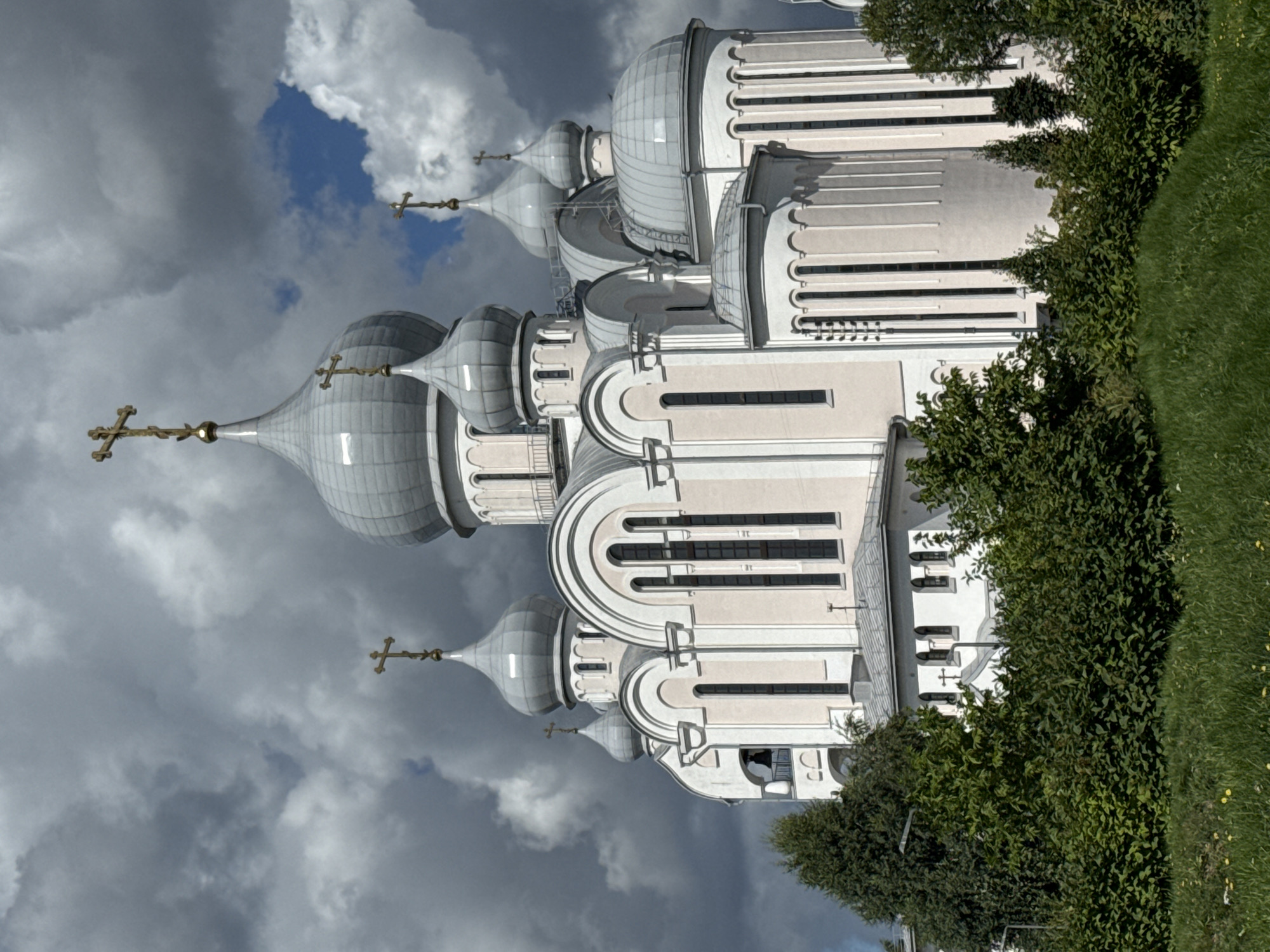 The image shows a large, ornate Orthodox church with multiple domes and crosses, set against a dramatic sky with dark clouds and patches of blue. The church is painted in light colors, possibly white and pale pink, with silver domes. The architecture features numerous arched windows and decorative elements. In the foreground, there is lush green vegetation, including trees and grass. The overall impression is one of grandeur and solemnity. This building is the Church of the Icon of the Mother of God of All Who Sorrow Joy in St. Petersburg, Russia.