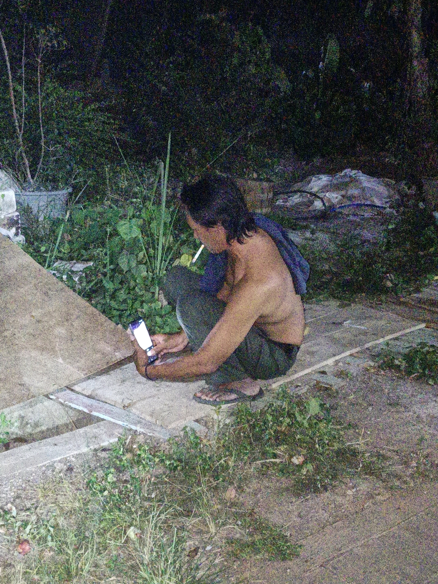 A shirtless man with long dark hair is squatting outdoors at night, holding a smartphone in his left hand and smoking a cigarette. He is wearing dark pants and sandals. He is positioned on a paved surface with dirt and overgrown vegetation surrounding him. In the background, there is dense foliage and some debris. The scene appears to be in a rural or semi-rural area.
