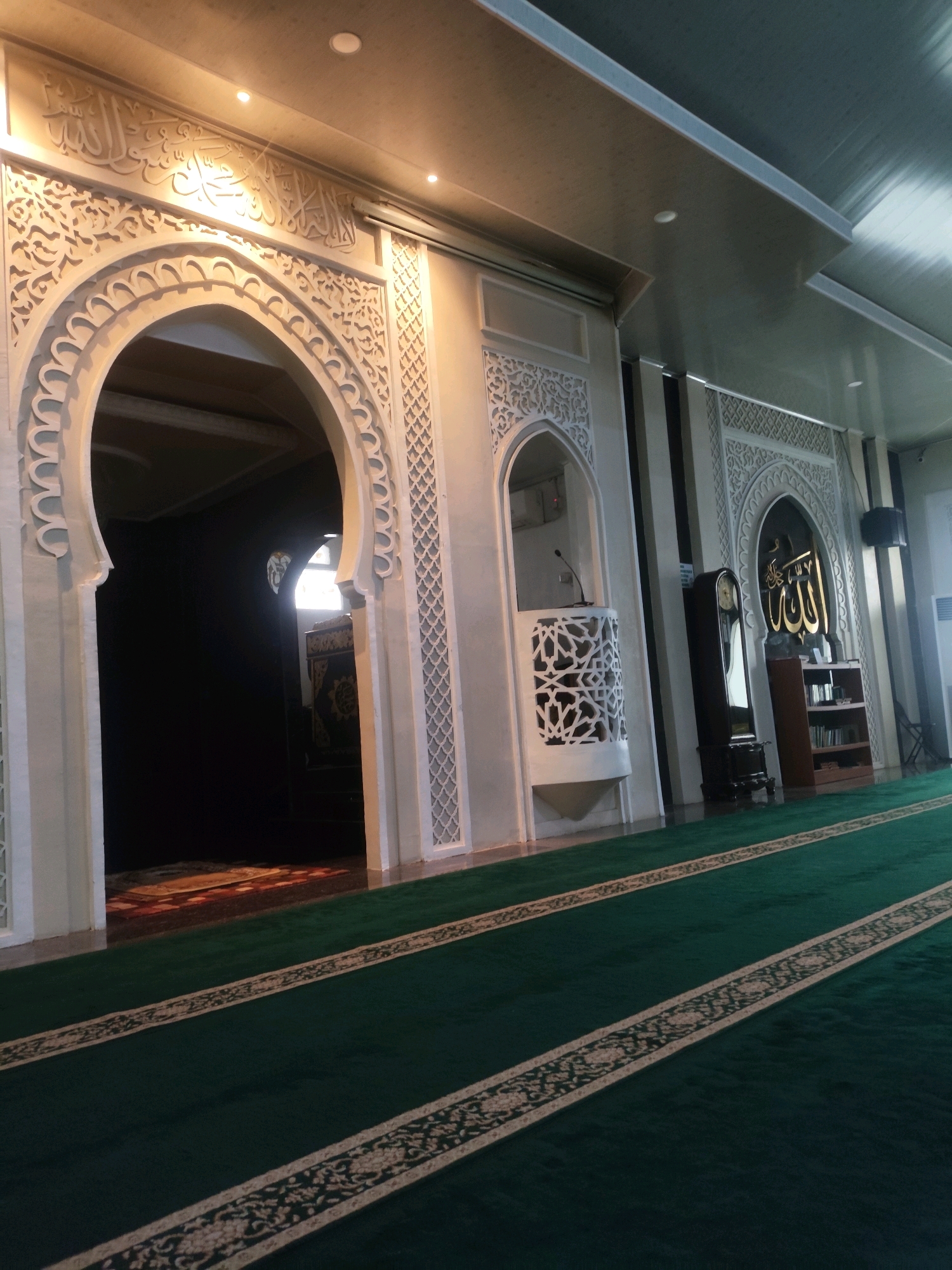 The image depicts the interior of a mosque, likely in Palembang, Indonesia, characterized by its Islamic architectural elements. The dominant feature in the foreground is a long, green carpet with intricate beige floral patterns running along its length, suggesting rows for prayer. To the left, a large, ornate archway with detailed carvings and Arabic calligraphy frames an entrance to a darker section of the mosque, where a prayer mat is visible. The archway is illuminated by spotlights from above, highlighting the detailed stucco work.
To the right of the archway, a patterned screen-like structure encloses a small pulpit, upon which a microphone is mounted. Further to the right, a tall, dark grandfather clock stands beside a wooden bookshelf filled with books. Above the bookshelf, an arched alcove features prominent Arabic calligraphy in gold against a black background. The walls are adorned with decorative panels that echo the Arabic calligraphy motif. The ceiling is a modern design with recessed lighting. The overall ambiance is one of reverence and architectural beauty, with a focus on prayer and Islamic tradition.