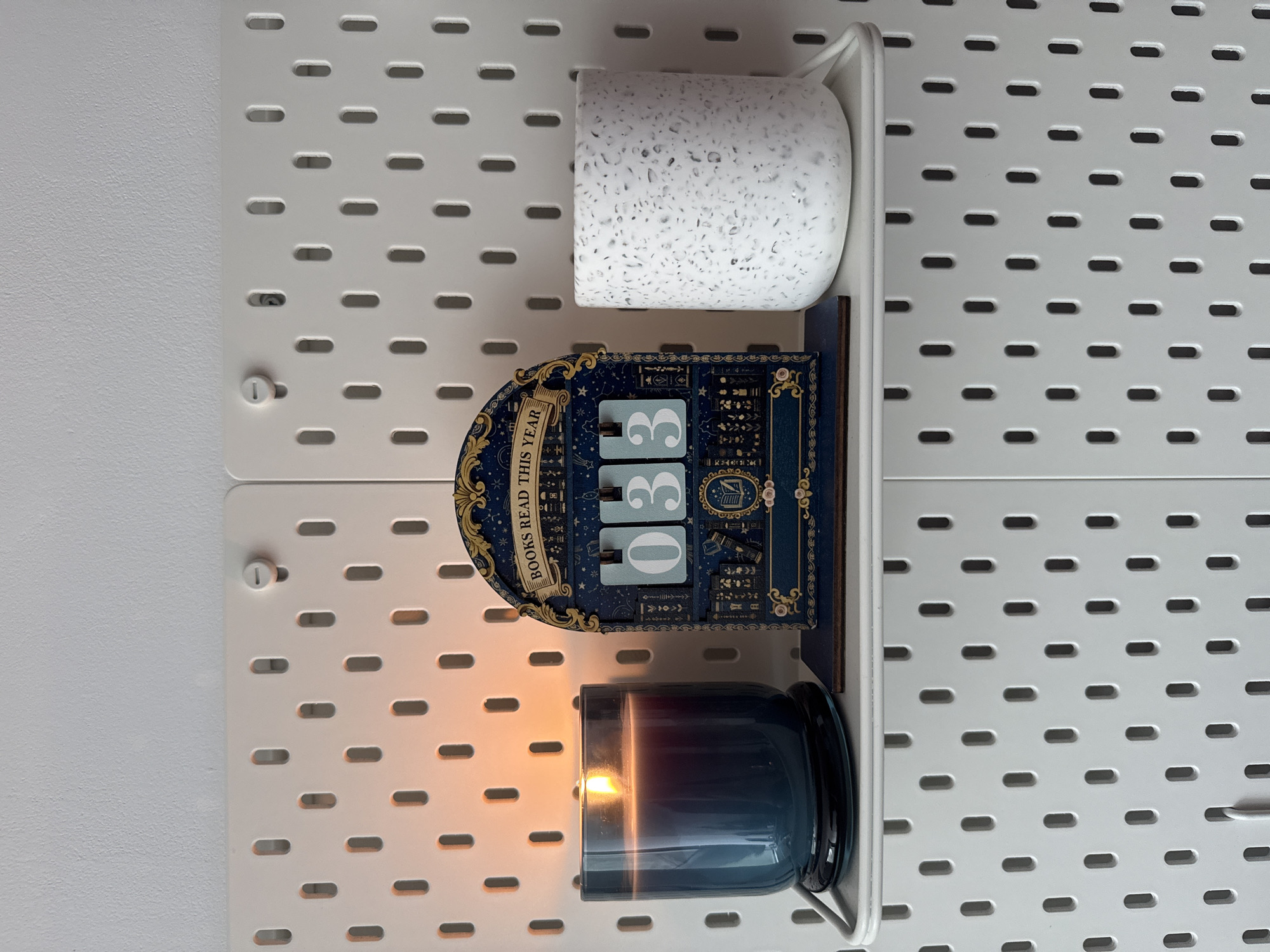 A neatly arranged indoor scene displays a white pegboard wall system, providing a backdrop for a white shelf holding three items. The pegboard itself is white with a repeating pattern of elongated oval holes, suggesting an organized storage solution, commonly found in residential settings in Riga, Latvia.
On the left side of the shelf, a dark blue candle in a glass jar is actively burning, emitting a warm, bright orange glow that illuminates the immediate area and reflects off the white surfaces. Centered on the shelf is a decorative, arched display piece, predominantly blue with gold accents. This object features the prominent text "BOOKS READ THIS YEAR" and displays the numbers "03," indicating a counter or tracker for books read. To the right, a white mug with a distinctive speckled pattern, resembling terrazzo with grey and black flecks, sits on the shelf.
The image is captured from a slightly elevated perspective, oriented vertically, emphasizing the organization and cozy atmosphere created by the lit candle. No people are visible, and the time of day is suggested to be evening or nighttime by the prominent glow of the candle.