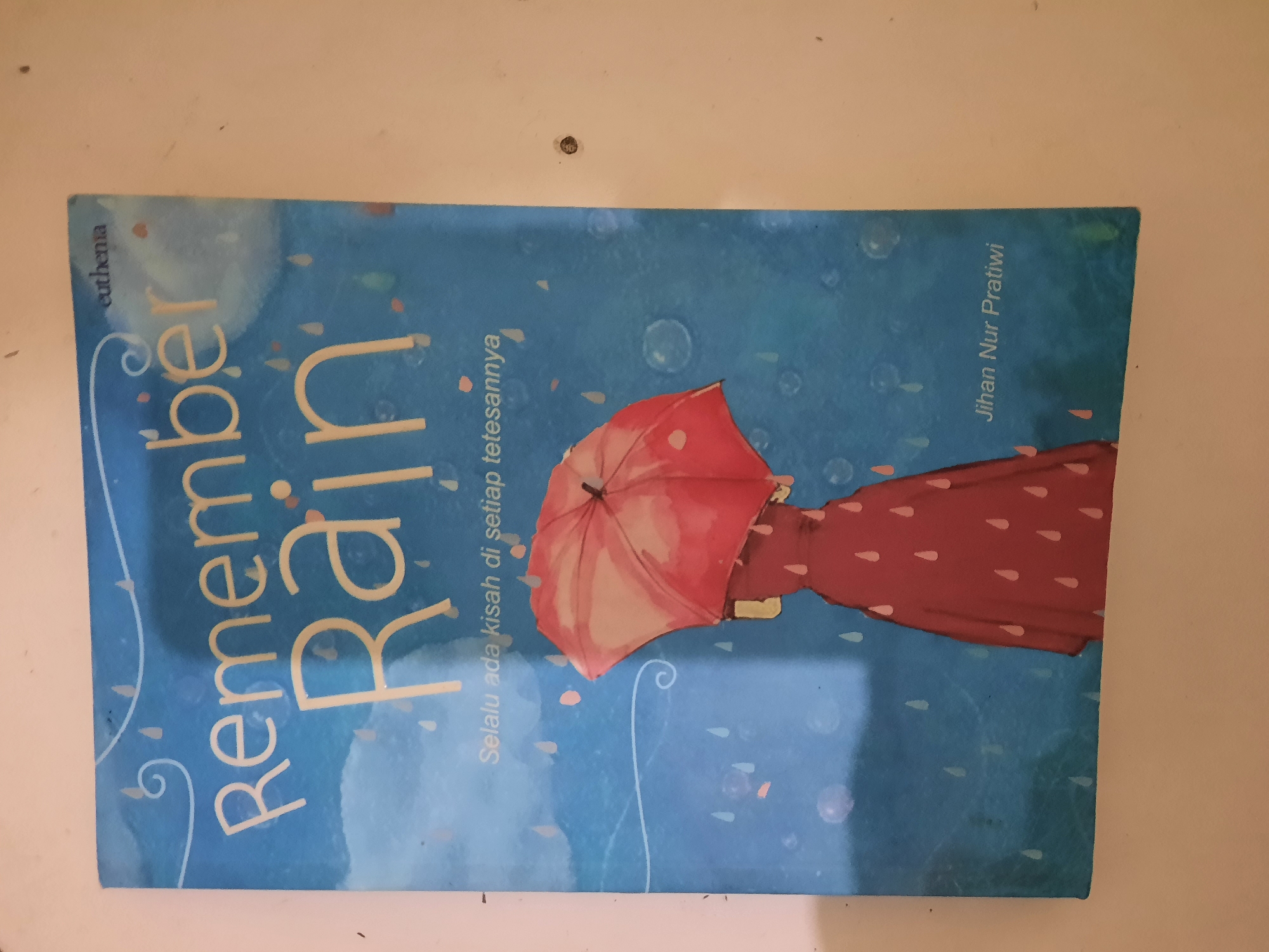The image is a close-up shot of a book cover titled "Remember Rain". The cover features a blue background with white stylized raindrops and bubbles. In the foreground, a figure in a red dress holds a pink umbrella. Below the title, there is a subtitle in Indonesian that reads "Selalu ada kisah di setiap tetesannya" (There is always a story in every drop). At the bottom right corner of the cover, the author's name, "Jihan Nur Pratiwi," is visible. The book is lying on a light-colored surface, possibly a table or wall, which appears to be a wall in Lhokseumawe, Indonesia.
