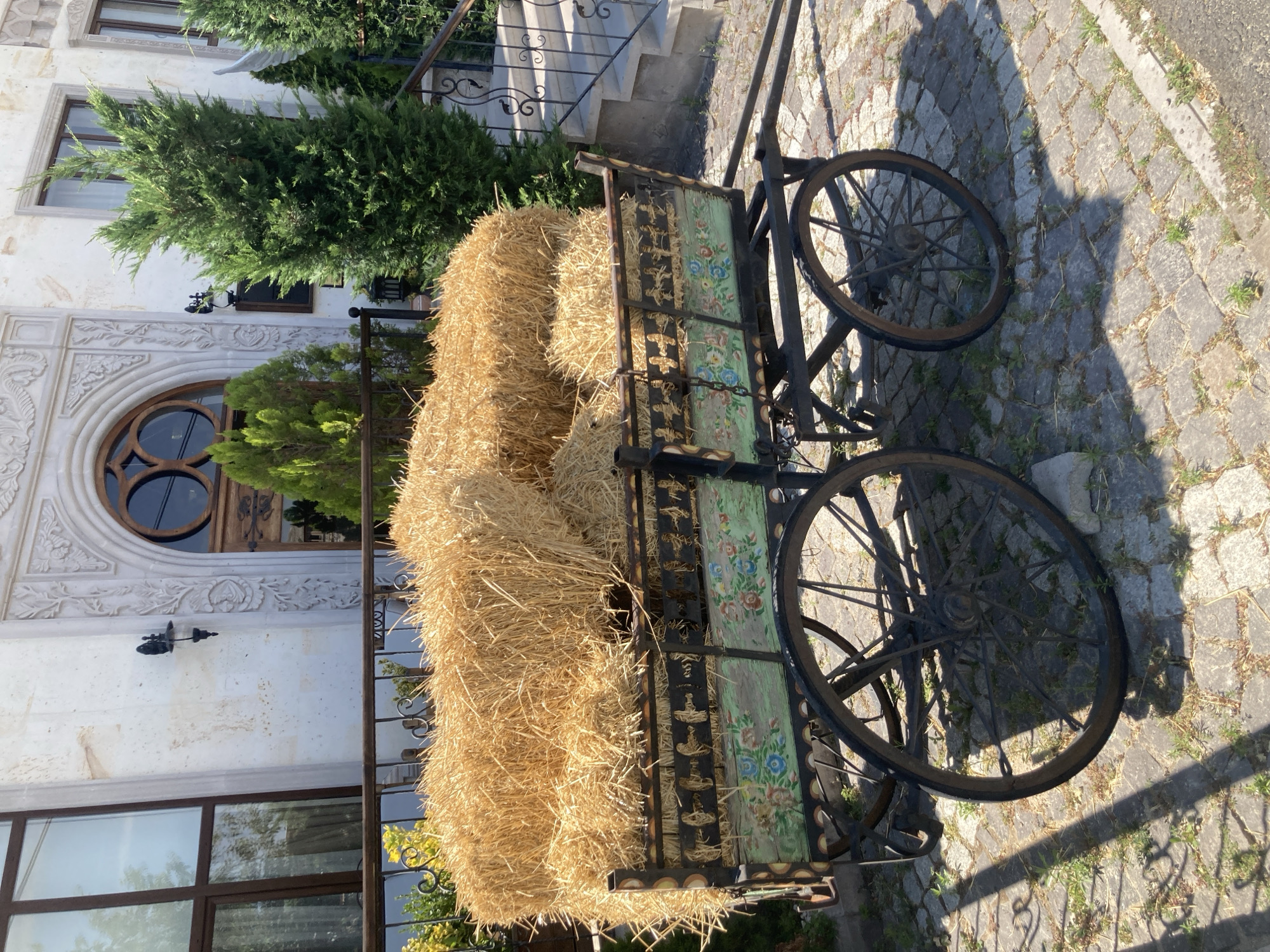 A rustic, horse-drawn cart filled with bales of golden straw is parked on a cobblestone path in front of a building. The cart is ornately decorated with painted floral designs and what appear to be silhouettes of horses and riders along its upper edge. The building in the background features light-colored stone with decorative arches and windows, and lush green cypress trees are planted around the entrance, suggesting a setting in Göreme, Turkey, a town known for its unique geological formations and cave dwellings. The scene is bathed in sunlight, casting shadows on the ground.