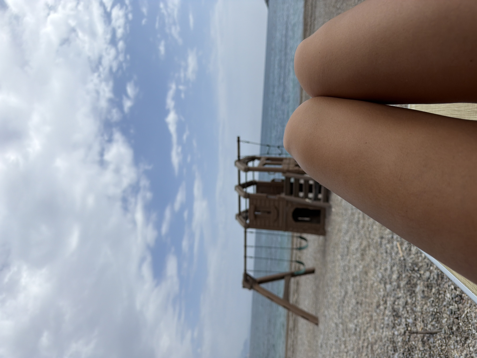 The image presents a scene at a beach in Prijevor, Montenegro. In the foreground, the tanned legs of a person sitting in a beach chair dominate the view. Beyond the legs, a children's playground structure with swings is visible on the beach. The shoreline meets a body of water, presumably the sea. The sky is overcast with a mix of blue sky and white clouds. The beach surface appears to be covered in pebbles.