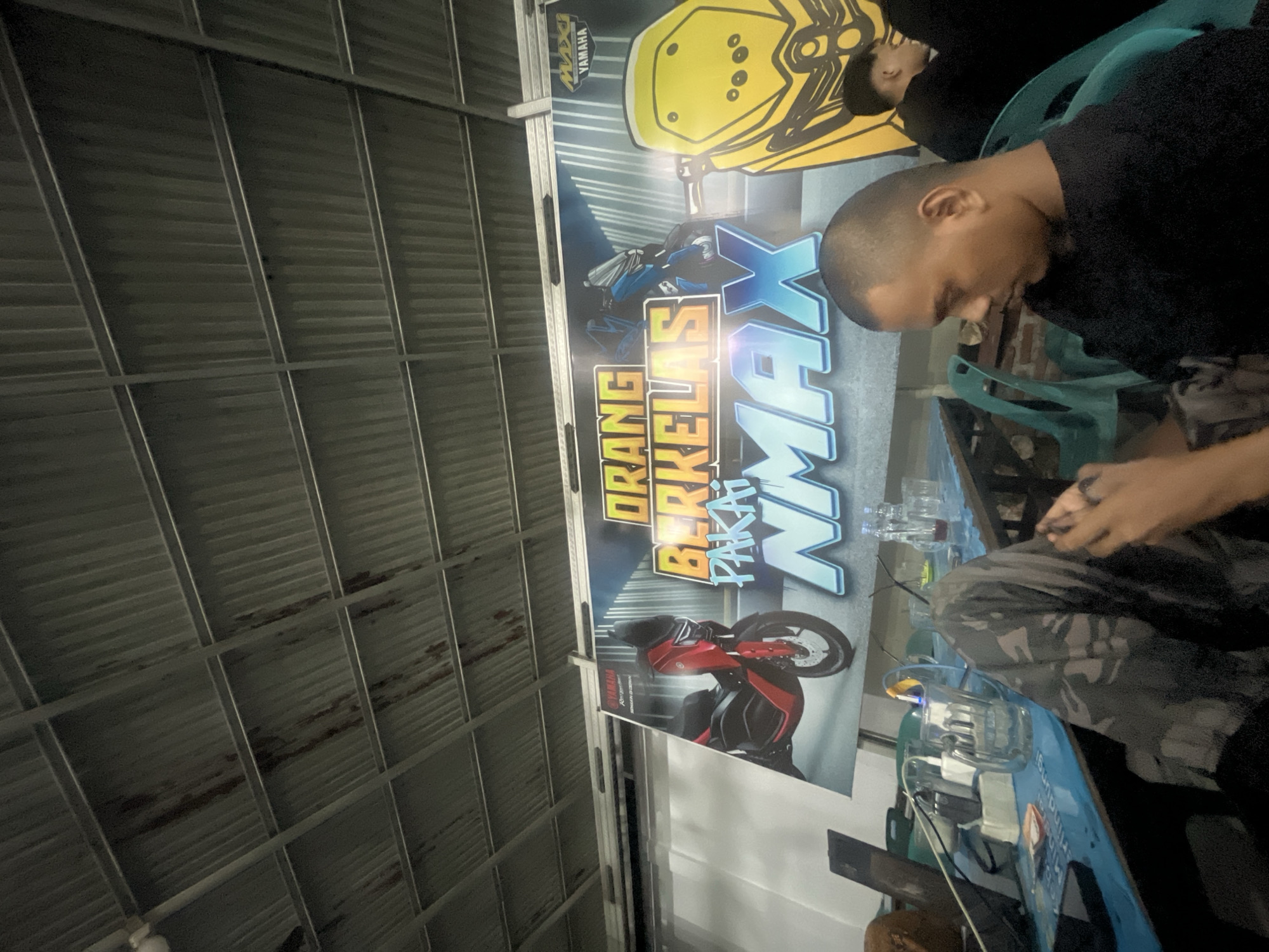 The image depicts a dimly lit interior space with a corrugated metal ceiling. In the foreground, a person wearing camouflage pants and a black t-shirt is seated and looking down at their phone. They are positioned at a blue table that displays various items including a laptop, power strip, glasses with liquid, and bottles. Behind them, a large banner is visible, featuring images of motorcycles and text that appears to be in Indonesian, possibly an advertisement. The banner showcases a red Yamaha NMAX motorcycle on the left and a yellow motorcycle part on the right. Other individuals are present in the background, partially obscured. The overall environment suggests an indoor gathering or hangout spot. While the image is labeled as being in Banda Aceh, Indonesia, specific identifying landmarks or details within the scene do not provide direct confirmation of this location.