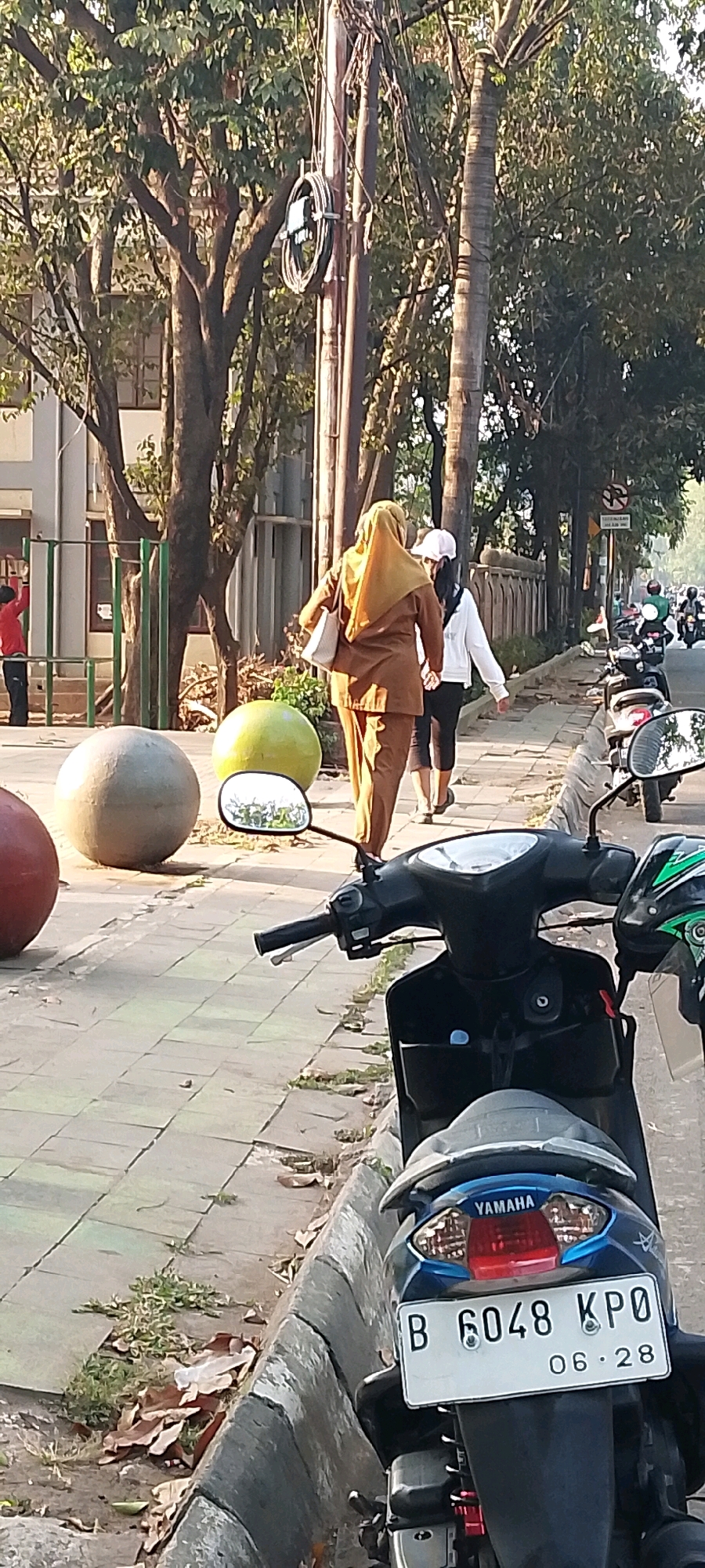 In Bekasi, Indonesia, two women are walking on a sidewalk next to a street lined with trees. The woman in the foreground is wearing a brown hijab and a matching brown outfit, carrying a white bag. The second woman is wearing a white jacket and black pants, with a white cap. A motorcycle is parked on the right side of the image in front of the sidewalk. In the background, several other motorcycles are on the street, with people riding them. The sidewalk has large, round decorative spheres on it. The area appears to be an outdoor public space with a building visible on the left.