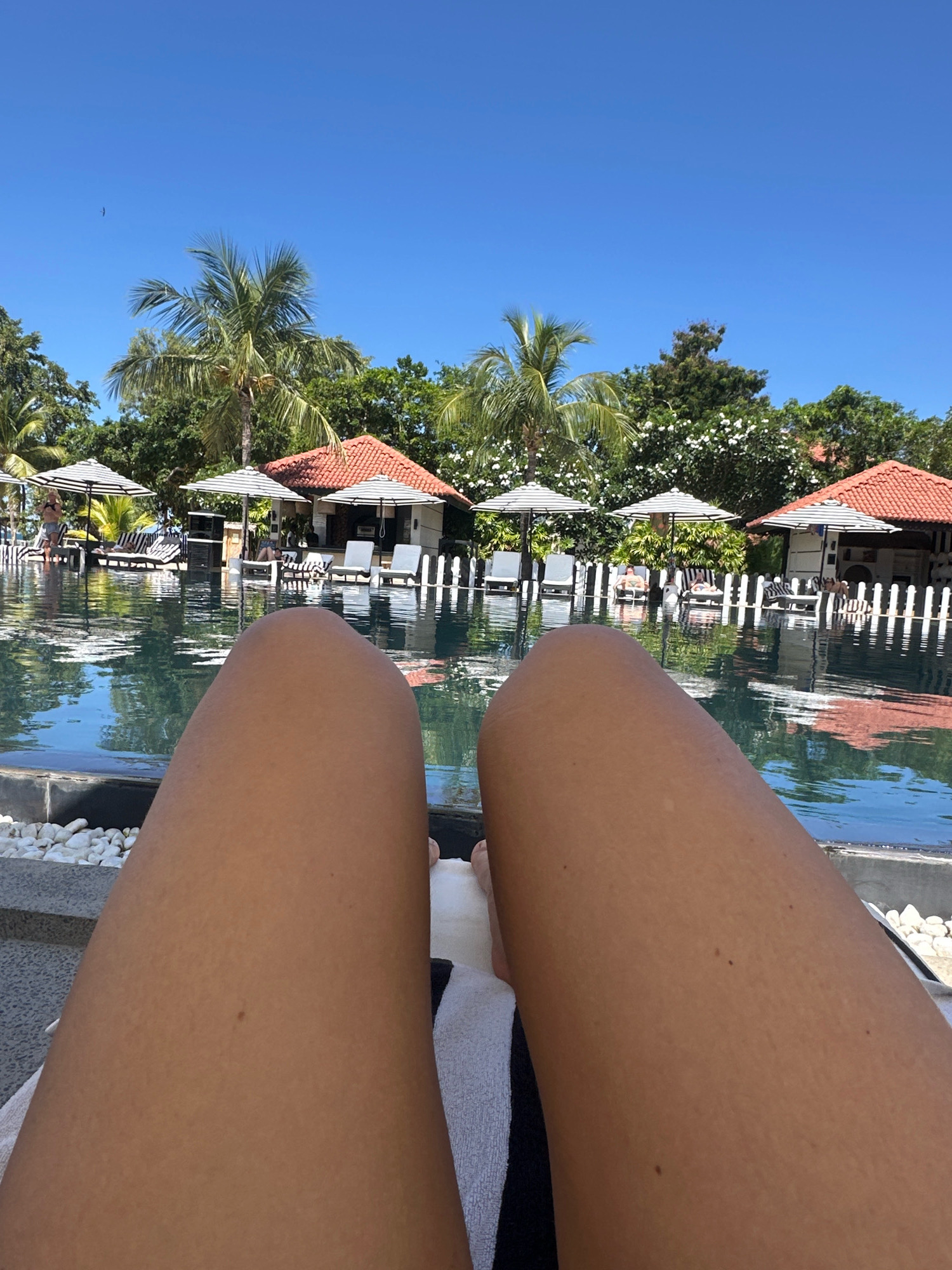 The image shows a first-person perspective of a person relaxing on a lounge chair by a swimming pool in Pasikudah, Sri Lanka. The person's legs are prominently in the foreground, with their tan skin visible. In the background, a resort setting unfolds with a clear blue sky overhead. A swimming pool with reflective water occupies the middle ground, bordered by white pebbles. Several lounge chairs with black and white striped umbrellas are arranged around the pool. Behind the pool, there are buildings with red tiled roofs, resembling cabanas or a restaurant. Lush green palm trees and other tropical foliage are visible, contributing to the resort ambiance. The overall scene depicts a vacation or leisure setting on a sunny day.