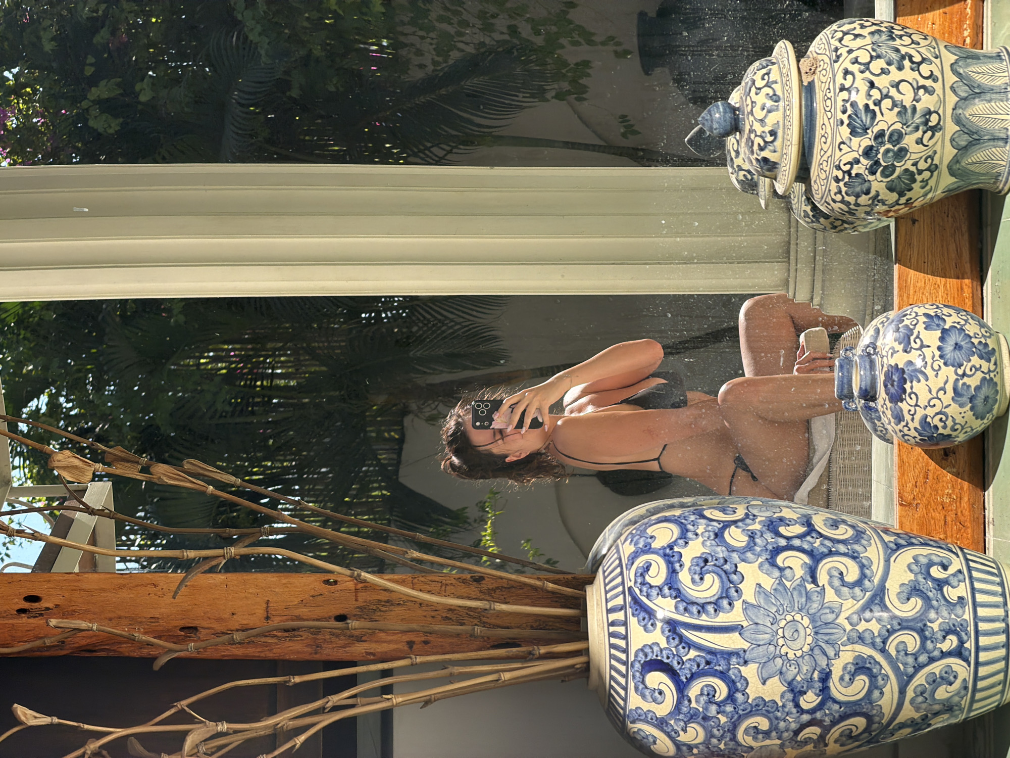 A woman is captured in a mirror or glass reflection, taking a selfie with a black smartphone that features white floral patterns on its case. She wears a dark bikini and appears to be seated or reclining, with her legs visible.
In the foreground, on a wooden surface, sit two large blue and white ceramic vases, intricately decorated with floral and scroll motifs. The vase on the right is tall and has a lid, while the one on the left is broader and more rounded. Part of a third, smaller blue and white pot is also visible. Dried branches or bamboo stalks are arranged in a holder, extending from the bottom left of the frame.
The reflection behind the woman and objects reveals a lush tropical outdoor environment with abundant green foliage, including palm trees. A white window frame runs horizontally across the mid-section of the reflected scene. The bright daylight suggests it is a sunny day. The setting combines an indoor space with a vibrant tropical vista, indicative of a location like North Kuta, Indonesia. There is no visible text in the image.