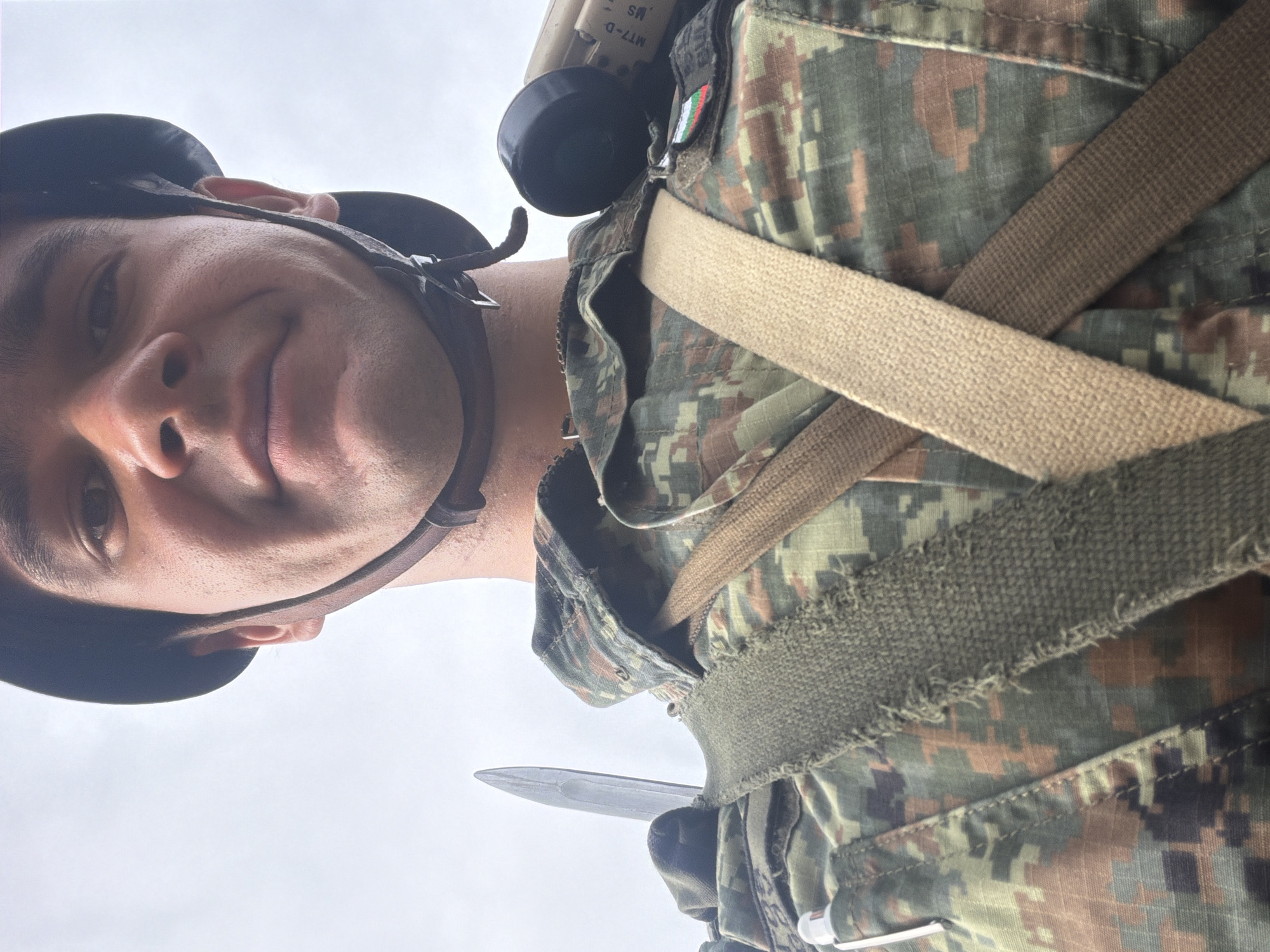 A close-up, waist-up selfie is captured outdoors against a bright, overcast sky. The subject is a young man wearing a military helmet and camouflage uniform. He has a bayonet attached to his uniform, with the blade visible over his left shoulder. On his right shoulder, a bandolier or strap is visible, and to the right of that, a device with a black circular end and a beige body with lettering "MT7-D MS" is attached. A patch with the Bulgarian flag (white, green, and red stripes) is visible on his uniform. The uniform itself is a pixelated camouflage pattern in shades of green, brown, and black. The lighting suggests daylight, possibly morning or late afternoon, with soft, diffused light. There are no discernible landmarks indicating the specific location within Veliko Tarnovo, Bulgaria.