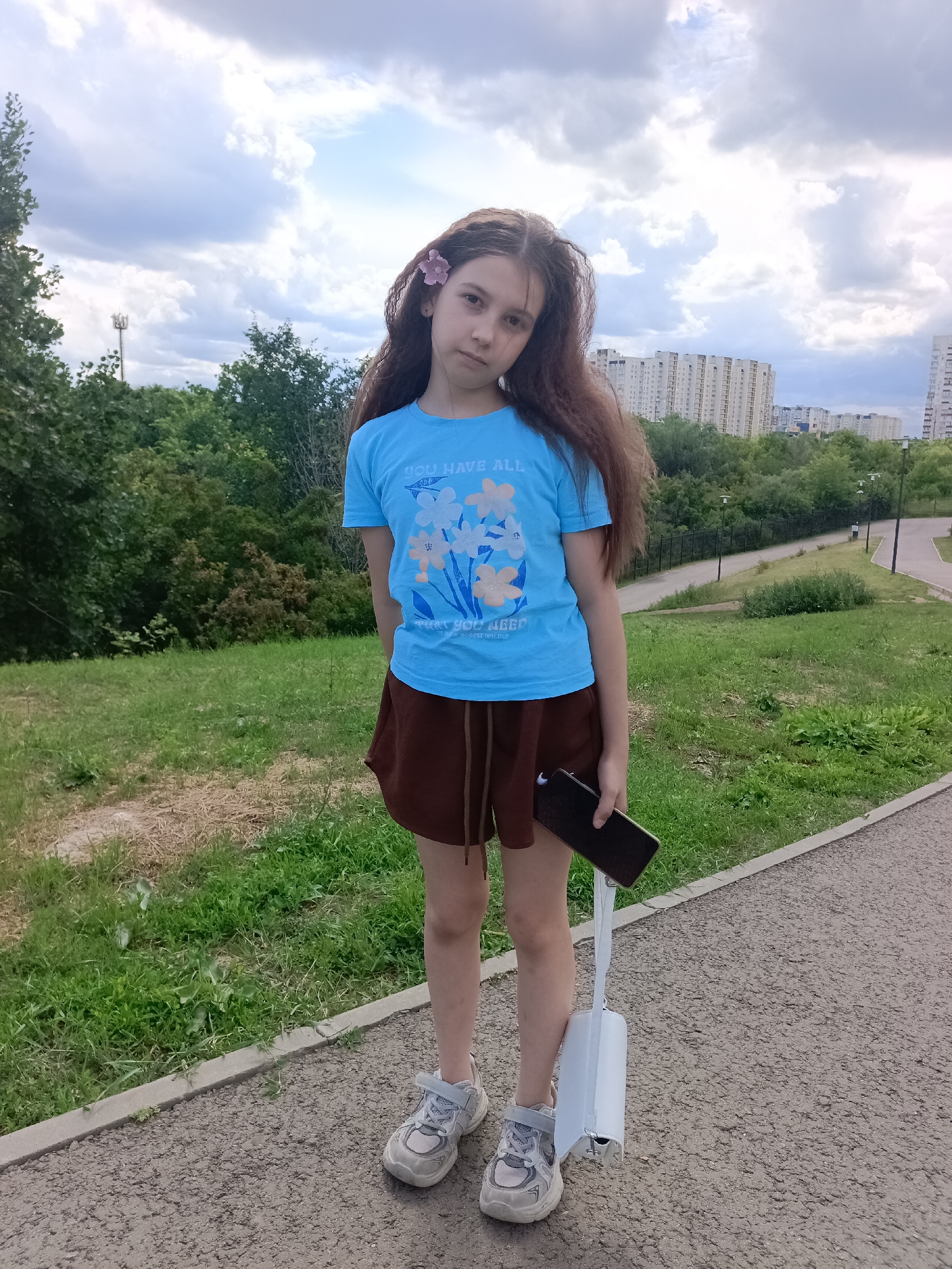 In this vertical, eye-level, full-shot photograph, a young girl stands on a paved path in an outdoor park. She is facing forward and looking directly at the camera with a neutral expression. The girl has long, dark brown hair that cascades down her back, and a small pink flower is tucked behind her right ear. She is wearing a bright blue t-shirt with a floral graphic and the text "YOU HAVE ALL THAT YOU NEED" and "TO BLOOM WILDLY" printed on it. Below her t-shirt, she wears a brown skirt. Her feet are clad in light gray sneakers.
In her right hand, she holds a dark-colored smartphone. A white shoulder bag with a rigid, rectangular body hangs from her left arm. The background features a grassy slope, a black metal fence, a winding paved path with lampposts, and a dense line of green trees. In the distance, several tall, multi-story apartment buildings are visible under a cloudy sky. The lighting suggests it is daytime, and the clouds indicate potentially overcast weather. The scene appears to be a typical urban park setting, possibly in Saratov, Russia, given the architectural style of the buildings.