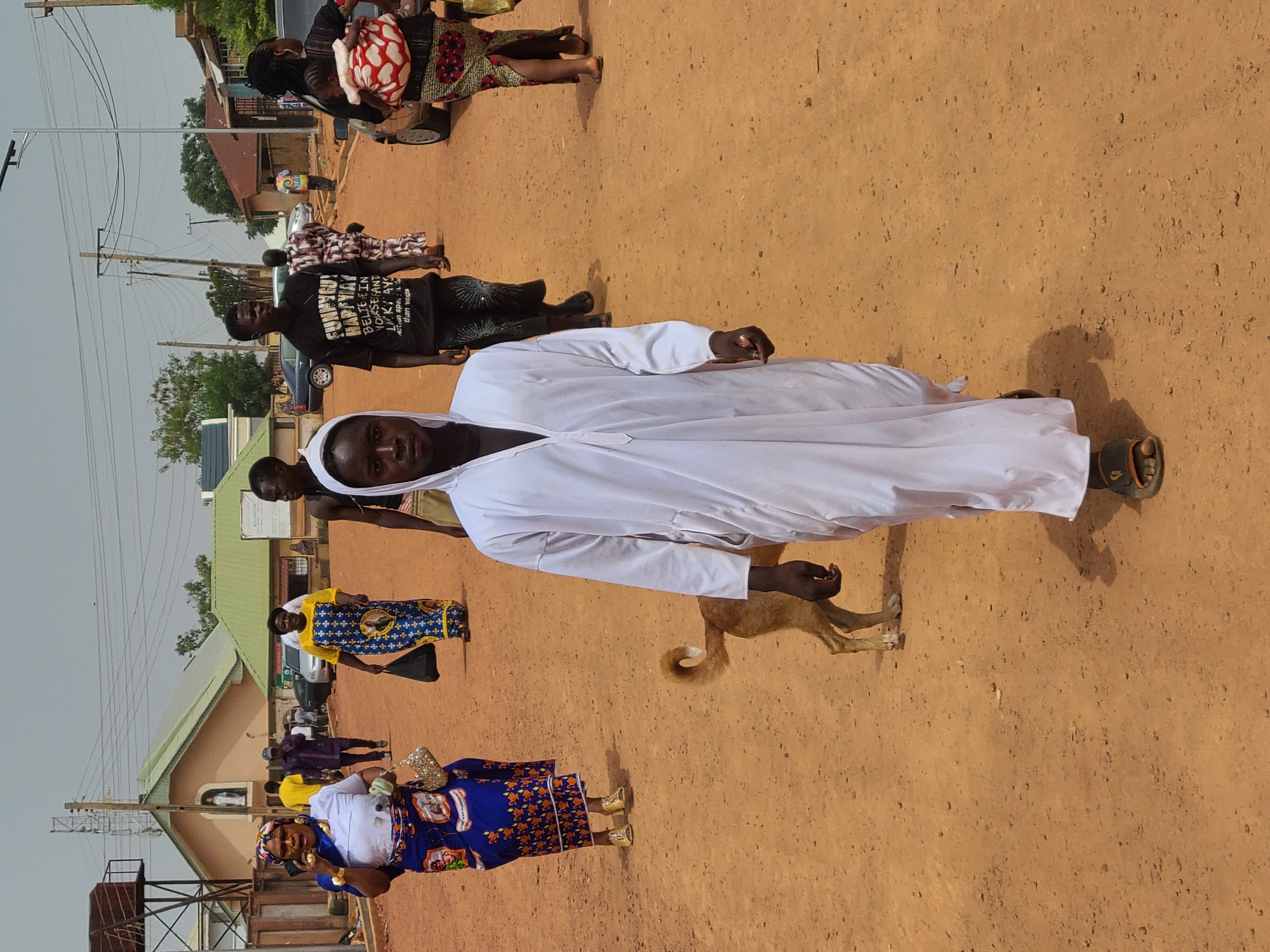 The image shows a street scene in what is most likely Jos, Nigeria. The ground is a reddish-brown dirt. A man in a white robe is in the foreground, facing the camera. Behind him and to the left, there is a brown dog and other people. Some are wearing colorful patterned clothing. There is a building with a light green roof visible in the background. A few vehicles are also present. Power lines stretch across the sky.
