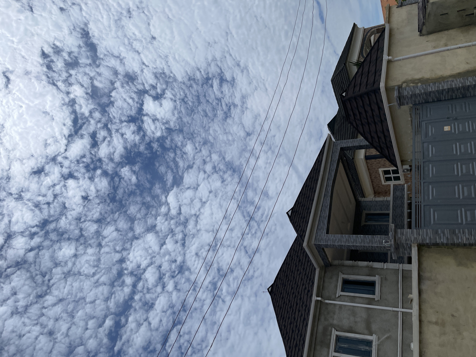 The image shows a residential street scene in Ado, Nigeria, under a sky filled with altocumulus clouds. The foreground features a portion of a residential building with a beige-colored exterior wall and a dark grey tiled roof. A grey metal gate is visible in front of the property, flanked by stone pillars. To the left, another part of a residential building is visible, with a grey stucco finish, white-trimmed windows, and a dark grey tiled roof. Power lines are strung across the upper portion of the image, running horizontally above the buildings. The architecture suggests a modern residential area.