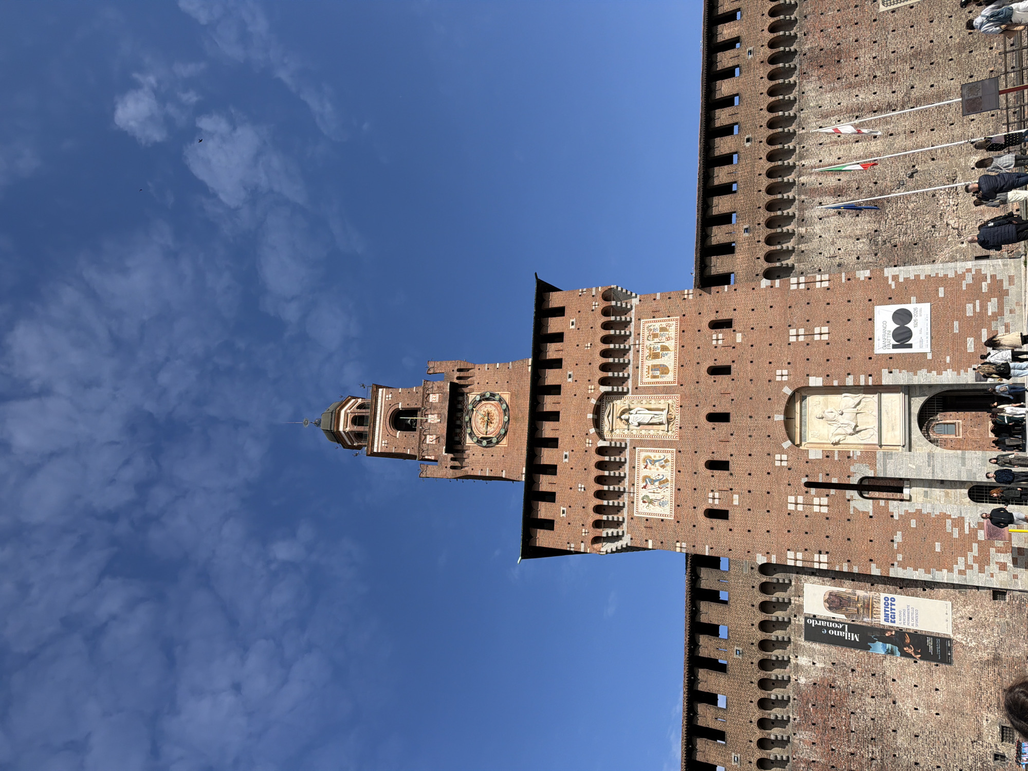 The image depicts the Sforza Castle in Milan, Italy. The view is from below, looking upwards at the castle tower, with a clear blue sky and some scattered clouds. The tower is brick, with architectural details such as arched windows, a clock, and decorative panels. On the right side of the image, the castle wall is visible, featuring battlements and flags. There are also advertisements displayed on the wall. Some people are visible near the base of the castle wall.