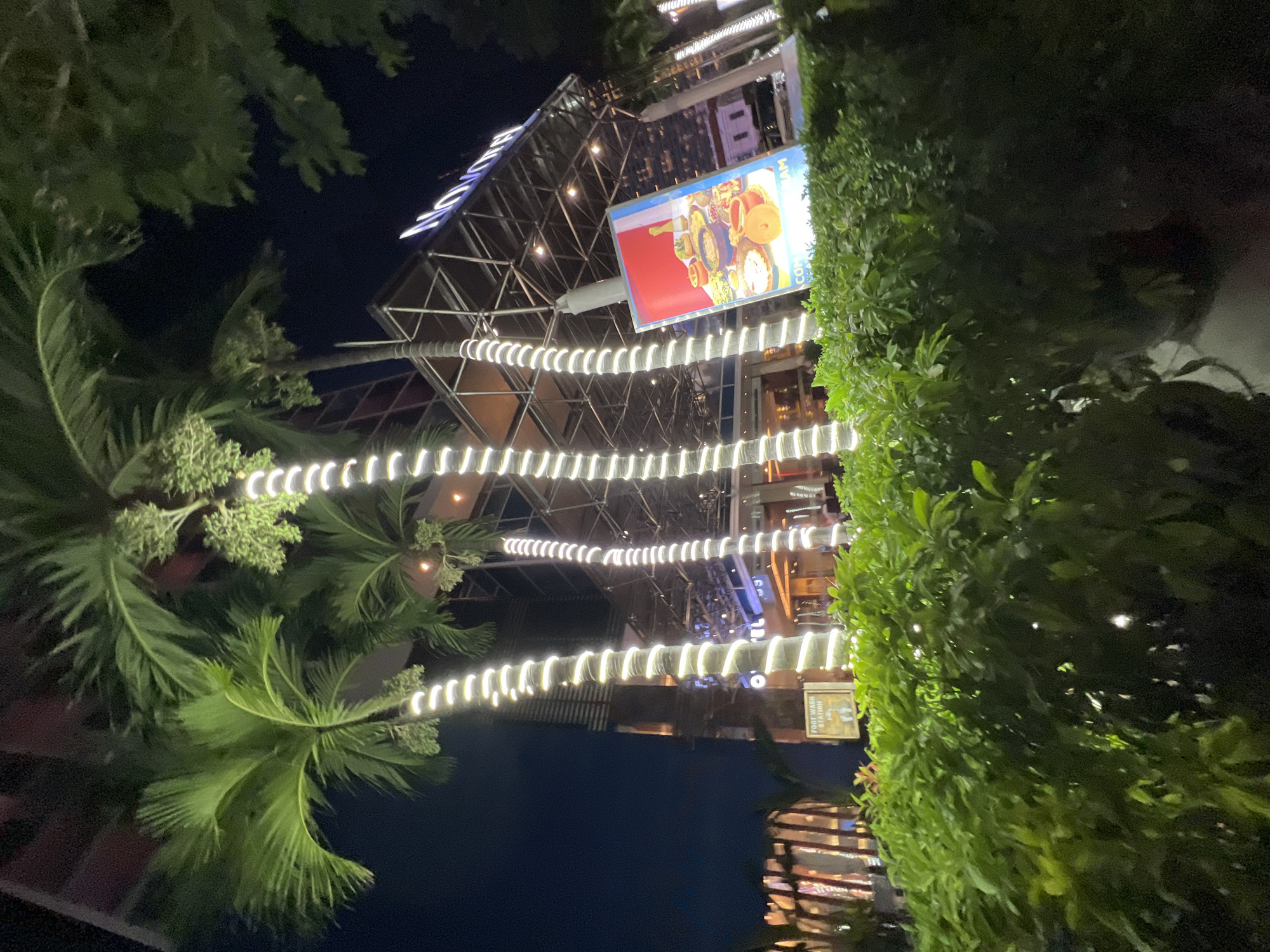 The image depicts the exterior of the Novotel Nha Trang hotel at night. The hotel's entrance is visible, with palm trees adorned with white string lights in the foreground. A large illuminated billboard displaying food items is positioned to the right, partially obscured by a dense hedge. Another sign, reading "FOOT WASH STATION," is visible on a pole behind the hedge. The building features a modern architectural design with glass facades and metal structures. The sky is dark, indicating nighttime. There are no people clearly visible in the frame.