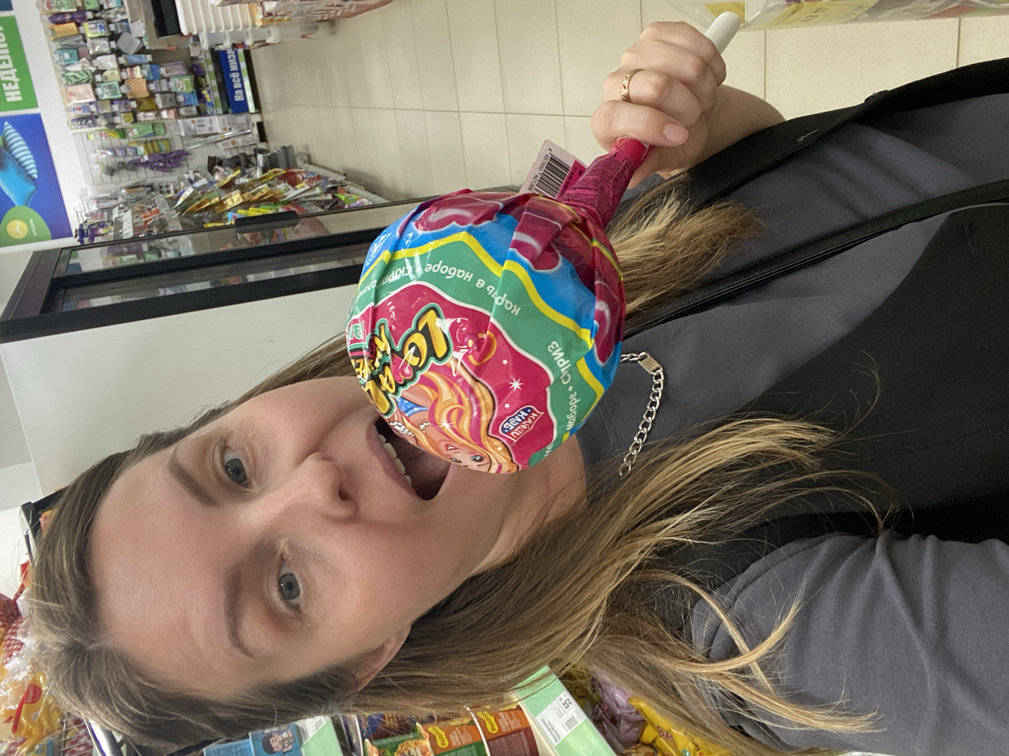 A woman with light brown hair is holding a large, colorful lollipop in front of her mouth, as if to eat it. She is wearing a dark grey t-shirt and a black vest. A silver chain necklace is visible around her neck. She is standing in a supermarket aisle, with shelves of products visible in the background. One shelf has a sign that reads "На всё низ" which is likely a price promotion. Another sign in the background shows a picture of pillows and the word "Неделя", indicating a weekly sale. The flooring is tiled. The scene appears to be in a typical retail environment.