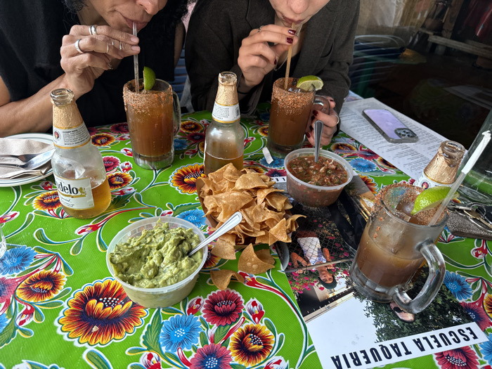 The image displays an overhead perspective of a table at Escuela Taqueria in Los Angeles, United States of America. The table is covered by a vivid green tablecloth patterned with large, colorful floral designs. Two individuals are partially visible at the top, both actively drinking from large, clear glass mugs. The individual on the left has rings on multiple fingers of their left hand and uses a clear plastic straw. The individual on the right has dark nail polish on their left hand and uses a wooden straw. Each mug contains a dark liquid, ice, a lime wedge, and a heavily seasoned rim. Three amber-colored Modelo beer bottles are present: one stands upright, one lies on its side, and one is inverted into a mug. Centrally, a clear plastic container of guacamole with a silver spoon, a brown paper-lined container of tortilla chips, and a clear plastic container of salsa with a silver spoon are arranged. A light purple smartphone with multiple camera lenses rests face-up on a white menu or advertisement, which also features a printed photograph of a person and the inverted text "ESCUELA TAQUERIA."