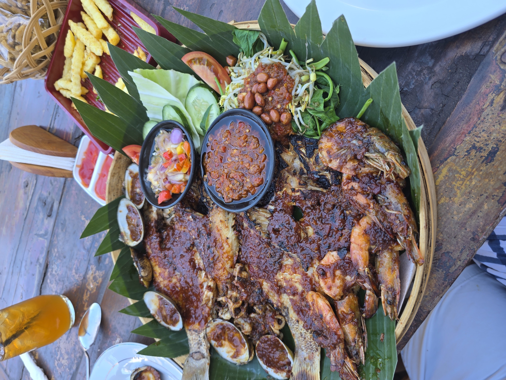 A large platter of grilled seafood, including two whole fish and several large shrimp, is presented on a bed of banana leaves. The seafood is coated in a dark, glossy sauce and garnished with clams, bean sprouts, peanuts, chopped tomatoes and onions, cucumber slices, and tomato wedges. Two small bowls of chili sauce and a side of french fries are also visible on the table. A glass of iced tea sits in the upper left corner of the image. The scene appears to be set outdoors on a wooden table, possibly at a restaurant in Seririt, Indonesia.