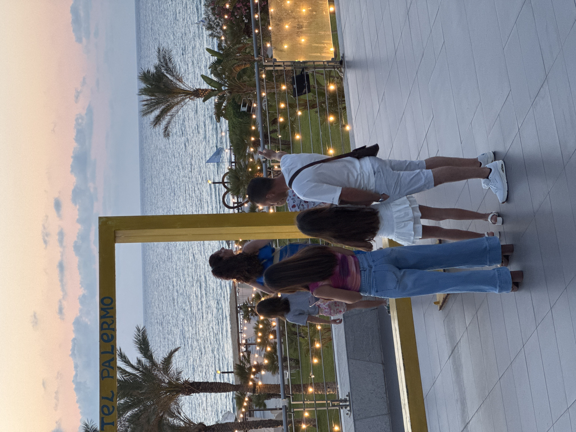 This media file captures a family or group of people posing for a photo within a large, yellow, rectangular frame, likely a prop. The scene is set outdoors in what appears to be a resort or beachfront area, indicated by the presence of palm trees and the sea. The time of day is dusk, as suggested by the soft, warm tones in the sky and the gentle illumination from string lights.
In the foreground, a man in a white t-shirt and light-colored shorts stands with his arm outstretched, seemingly directing the photo. He wears a dark crossbody bag. To his right, a woman in a white dress and sandals is visible. Behind them, three individuals, possibly children or younger adults, are also positioned within the frame. One person wears a pink and blue tie-dye shirt and blue jeans. Another is in a blue and yellow striped top, and the third person in the middle has long, dark hair.
The setting features a paved walkway made of light-colored square tiles. Beyond the walkway, the calm sea stretches towards the horizon. A metal railing with decorative elements separates the walkway from the area below, where more palm trees and greenery are visible. The string lights are draped along the railing and in the background, adding a festive ambiance to the scene. The overall mood appears relaxed and joyful, characteristic of a vacation or a special gathering.
Visible text on the yellow frame reads "TEL PALERMO," possibly indicating a hotel or a local establishment related to Palermo. The weather seems pleasant and calm, with a soft twilight glow.