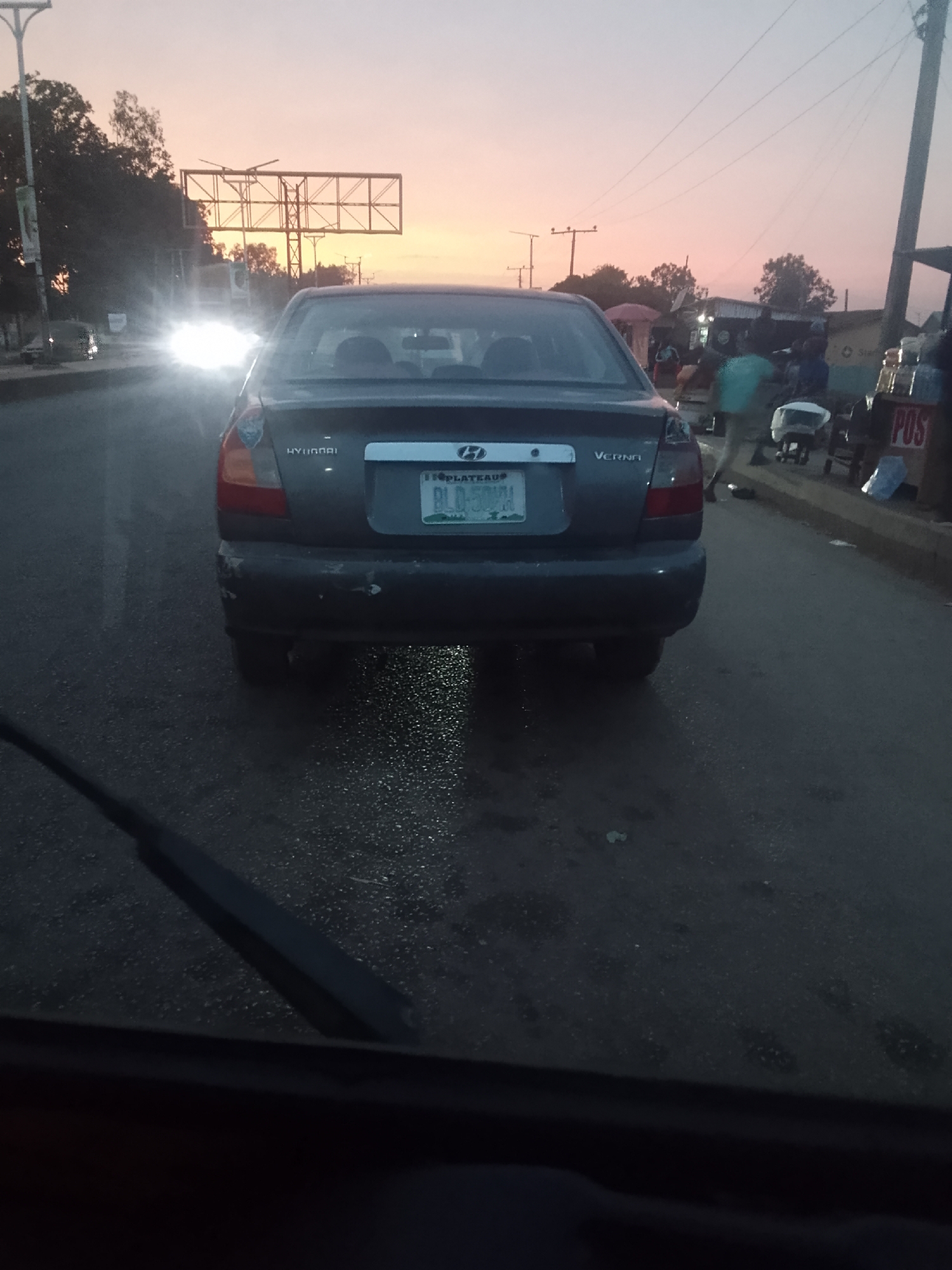 The image depicts the rear view of a dark gray Hyundai Verna car with license plate "BLD 505 LH" as seen from the windshield of another vehicle. The car is positioned on a wet asphalt road with a street with stalls and people on the right side. The scene is in Bukuru, Nigeria, suggested by the license plate. The time appears to be dusk or dawn, indicated by the soft, muted light and the pink-tinged sky. In the distance, traffic lights are visible, and a light source comes from the opposite direction.