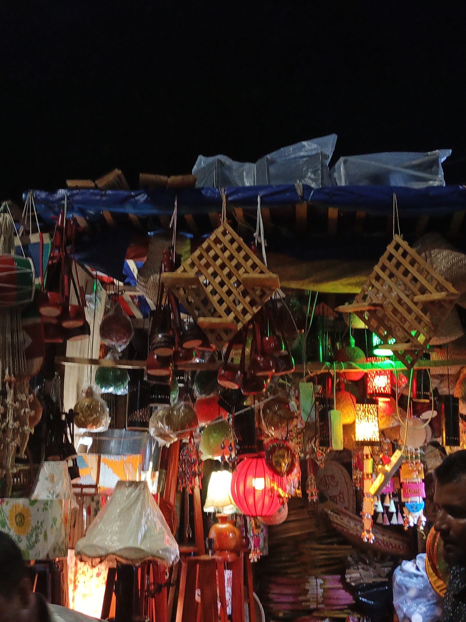 The image depicts a bustling market scene in Dhaka, Bangladesh at night. Numerous handcrafted lanterns and hanging decorations are displayed under a tarpaulin shelter. The lanterns are of varying shapes, colors, and materials, some illuminated by internal lights. A man's face is visible in the lower right corner, partially obscured. Other individuals are partially visible in the lower left corner. The background is dark, suggesting a nighttime setting.