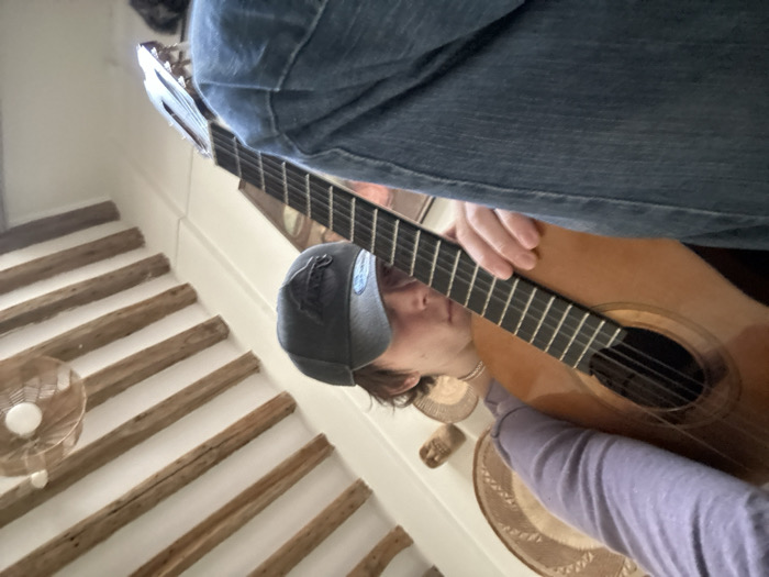 An individual is depicted indoors, seated, with a light brown classical or acoustic guitar across their lap. The person wears a light purple long-sleeved shirt, blue denim jeans, and a dark grey baseball cap with an emblem on the side. Their head rests against the body of the guitar, and one hand is positioned on the guitar's fretboard. The room's ceiling features prominent exposed dark wooden beams against a white background. Several woven, basket-like decorative objects are mounted on the wall behind the individual, and a light fixture with a woven shade hangs from the ceiling to the right. The architectural style, specifically the exposed ceiling beams, is characteristic of older buildings found within Paris, France.