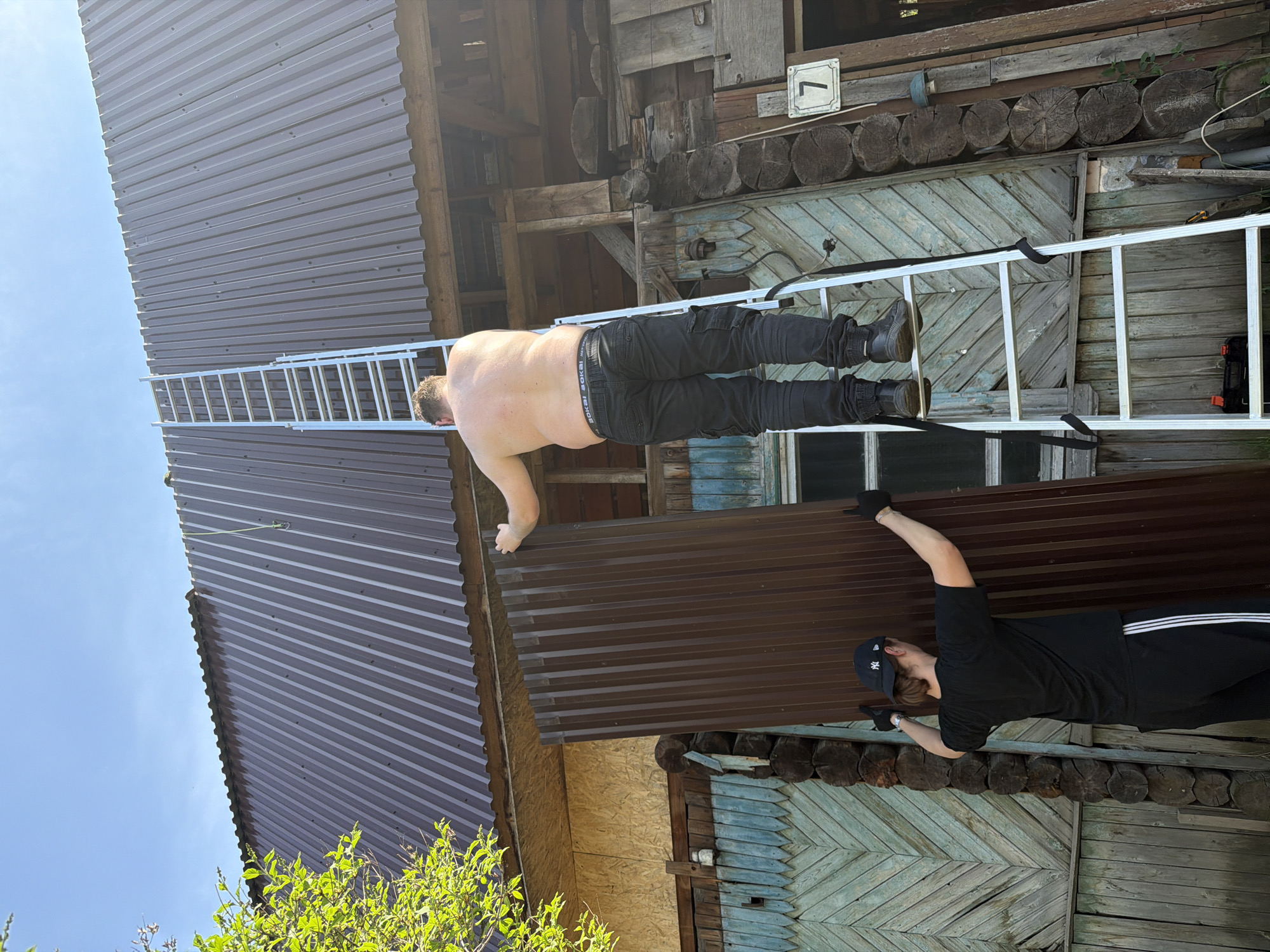 A photograph shows two men on a ladder next to a building. One man stands at the base of the ladder wearing a black t-shirt, black pants with white stripes, a black baseball cap, and gloves. The other man is positioned higher up on the ladder, shirtless with black pants and boots. He reaches towards the roof of the building. The building has sections of brown corrugated metal and weathered wooden walls. The background features a blue sky and greenery. A small sign with the number "7" is visible on the wall. The location is likely Perm, Russia, as it resembles architecture typical of the region.