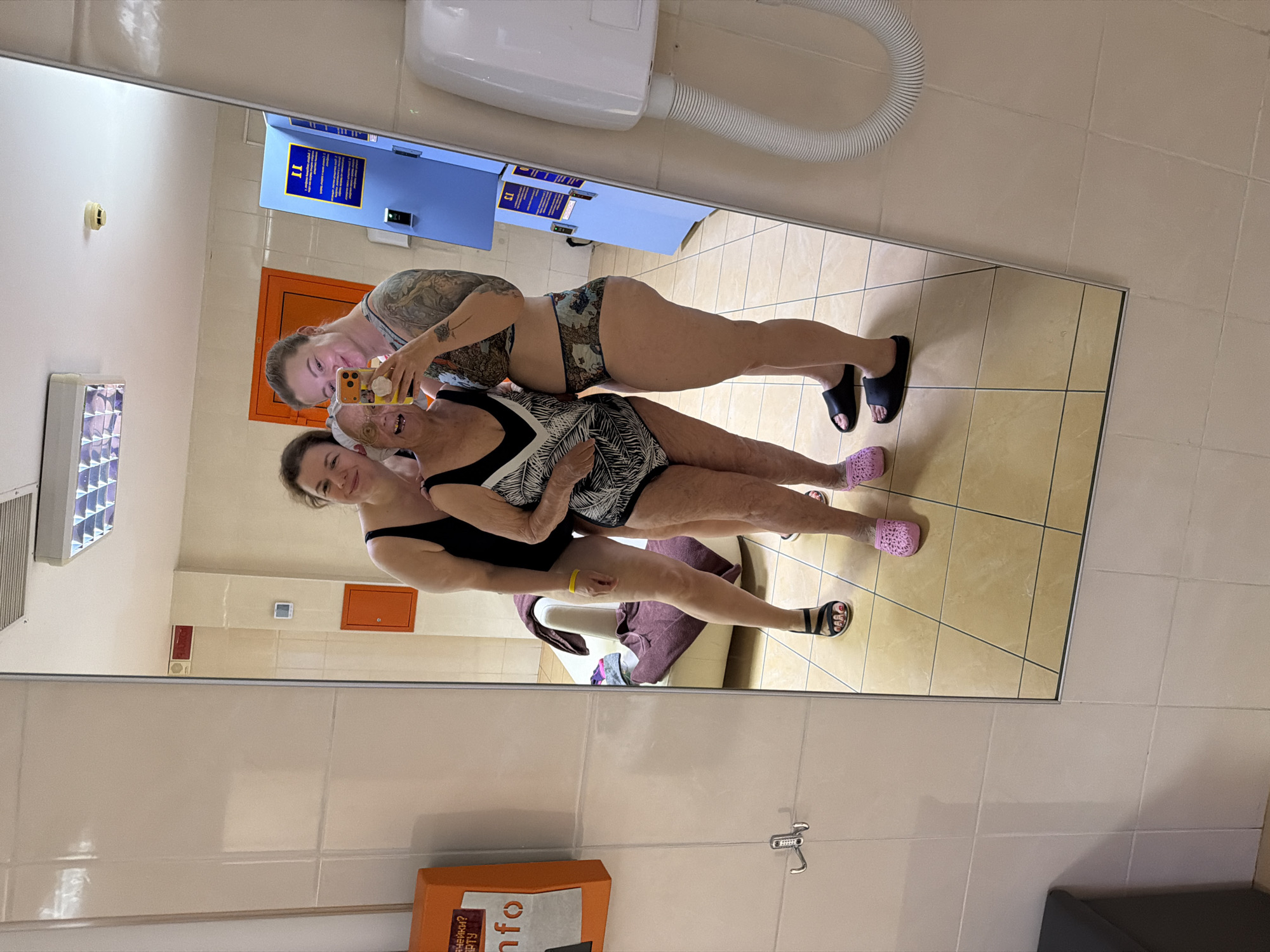 In Ждановіцкі сельскі Савет, Belarus, a mirror reflects a scene of three women posing closely for a selfie. The woman at the top, with visible tattoos on her arm, holds a phone to capture the image. The other two women stand below her, embraced. All three are wearing swimwear and footwear suitable for a pool or sauna environment. Behind them, a bench with towels sits within the tiled room, suggesting a changing area or spa. Lockers and a hand dryer are mounted on the wall. The scene is well-lit, and the reflection showcases the tile floor and wall details of the room.