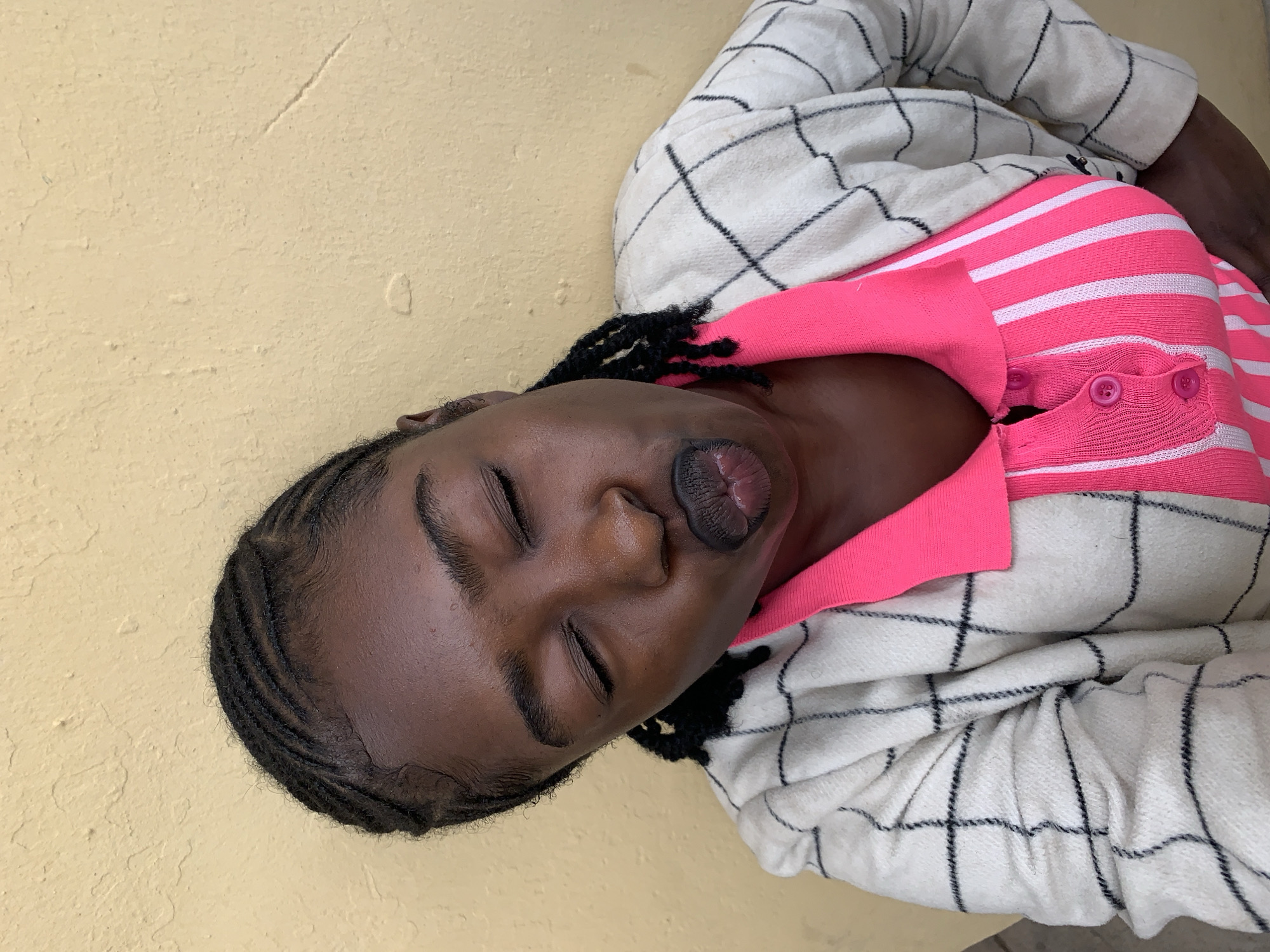 A close-up portrait, presumably taken in Rafin Par, Nigeria, features a female individual against a pale yellow, textured wall. The subject's head is tilted to the viewer's right, with her eyes closed and lips deeply pursed into a puckered expression. She has dark skin and wears dark lipstick. Her hair is styled in close-set cornrows across the scalp, with several longer, loose braids visible near her ears and shoulders. She is dressed in a vibrant, collared pink top with vertical white stripes and three visible pink buttons on the placket. Over this, she wears an off-white or cream-colored, long-sleeved jacket or cardigan featuring a black grid or plaid pattern, made from a visibly textured fabric. The wall behind her displays surface irregularities, minor cracks, and areas of worn texture, consistent with an aged exterior surface.