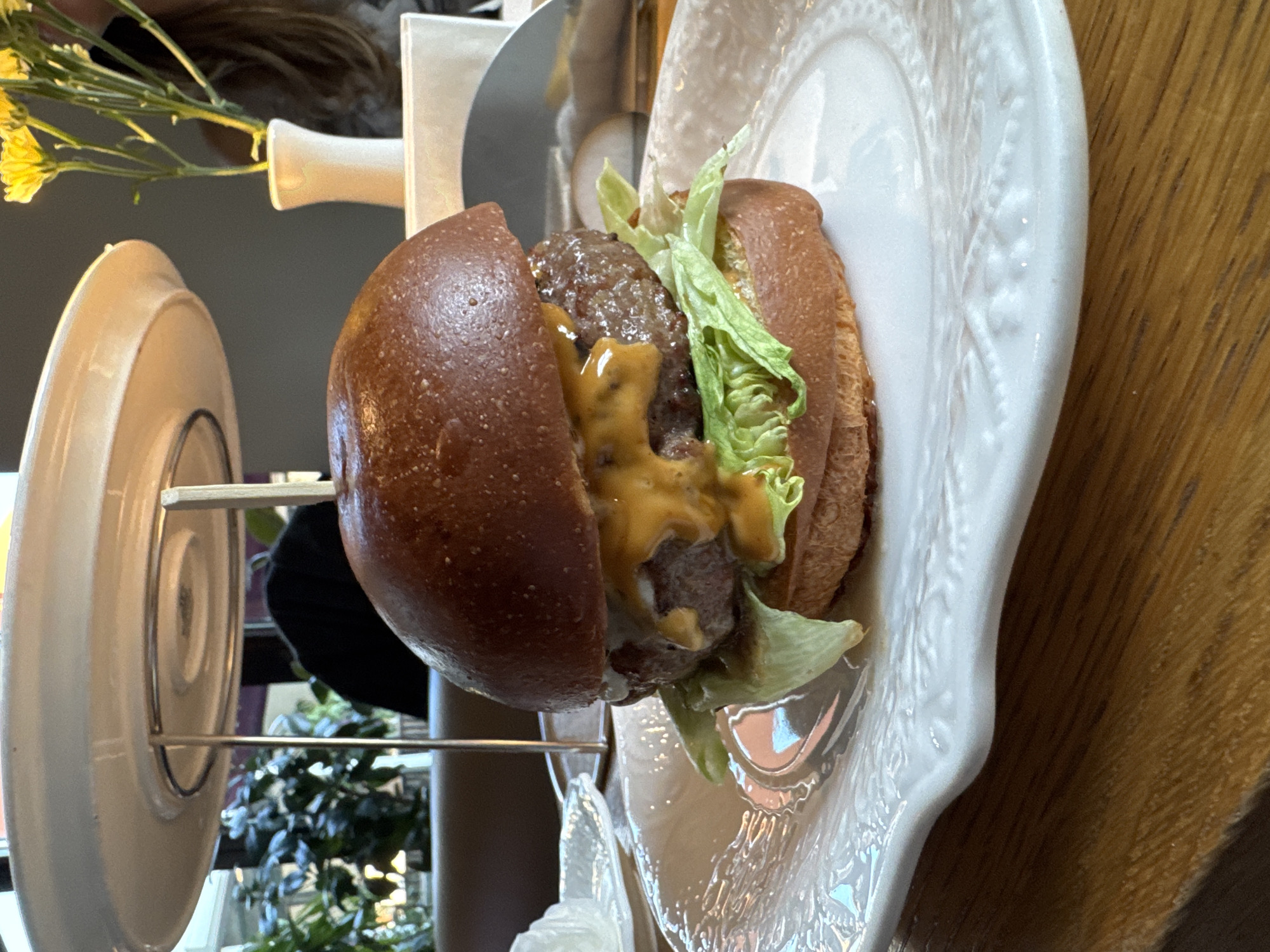 The media file displays a close-up, portrait-oriented shot of a single burger, presented in an indoor setting. The burger, positioned centrally, is held vertically by a metal skewer that extends through its layers and is supported by a white, angular stand. It features a dark, glossy bun, a thick meat patty, melted yellow cheese sauce drizzled over the patty, and fresh green lettuce.
The burger rests on a white, ornately detailed plate with a scalloped edge, which is placed on a warm-toned wooden surface, likely a table. Reflections are visible on the glossy surface of the plate, and the texture of the wood grain is prominent.
In the blurred background to the left, a small bouquet of yellow flowers in a white vase is visible. Further back and to the right, soft green foliage of plants can be discerned, suggesting an indoor environment like a restaurant or cafe. A blurred human head or hair is faintly visible behind the flowers.
The lighting appears natural, suggesting daytime. No discernible text or specific location cues are visible within the image itself, though the context provided indicates the location is Moscow, Russia. The overall scene focuses on the appetizing presentation of the burger.
