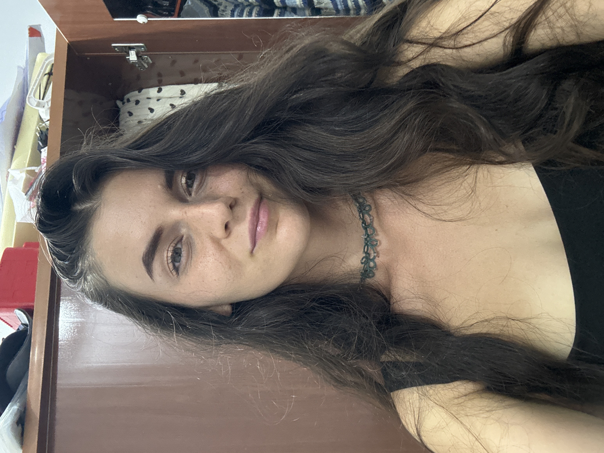 A brunette woman with fair skin and freckles is posing indoors, lying on her side with her head close to a wooden cabinet. She is wearing a black top and a dark green choker necklace. Her long, wavy hair is spread around her. The background shows the interior of a cabinet with items inside, and what appears to be a partially visible room with other furniture. There is no indication that this photo was taken in Mũi Né, Vietnam.