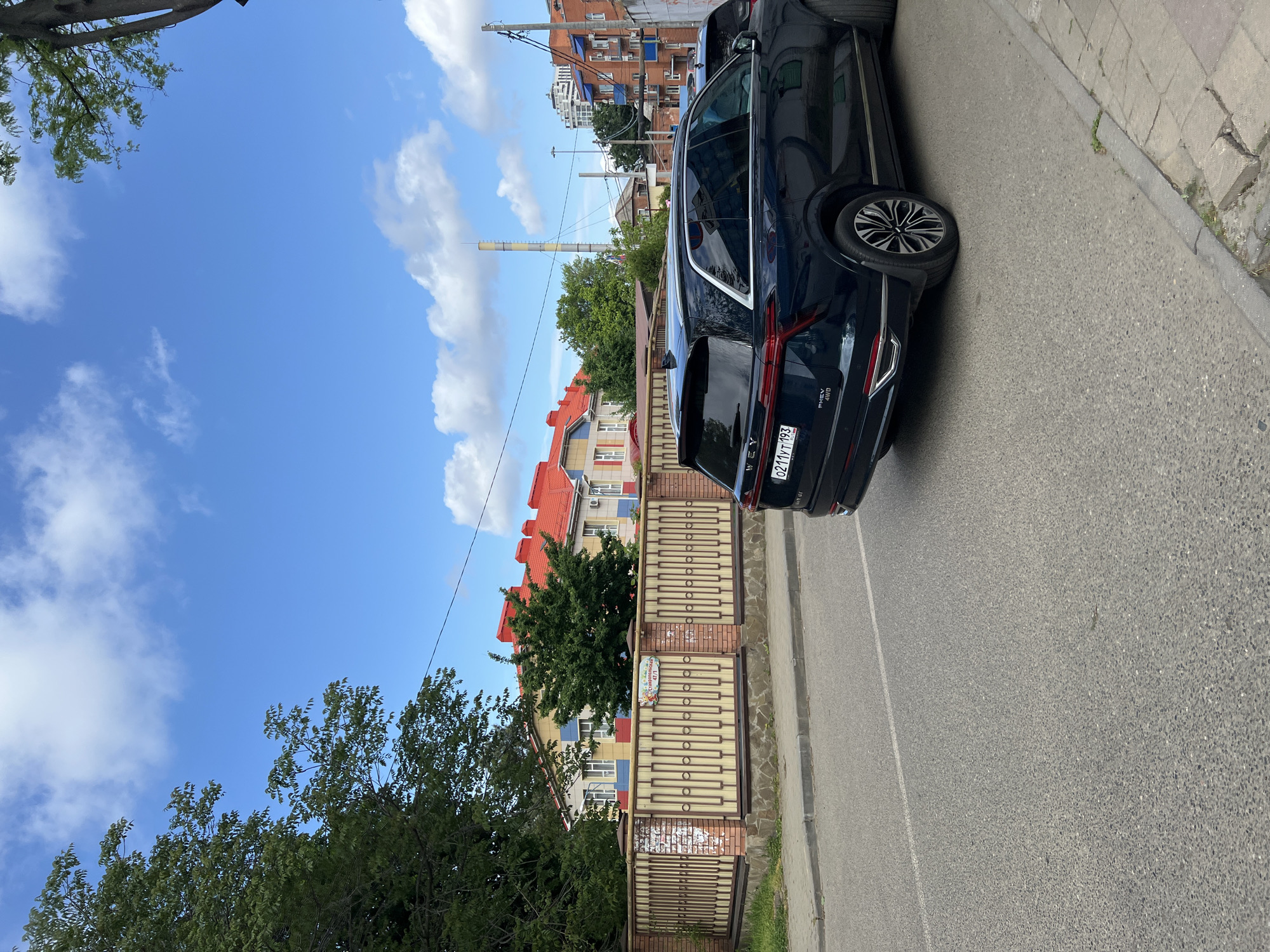 The image shows a dark blue WEY PHEV 4WD parked on a city street in Krasnodar, Russia. The license plate reads "O211YT193". Behind the car is a fenced building with a sign indicating "Детский сад 171" (Kindergarten 171). The building has a red roof and a yellow and blue facade. A tall industrial smokestack is visible in the background. The sky is blue with scattered white clouds. There are trees lining the street, casting shadows. The pavement is gray and cracked.
