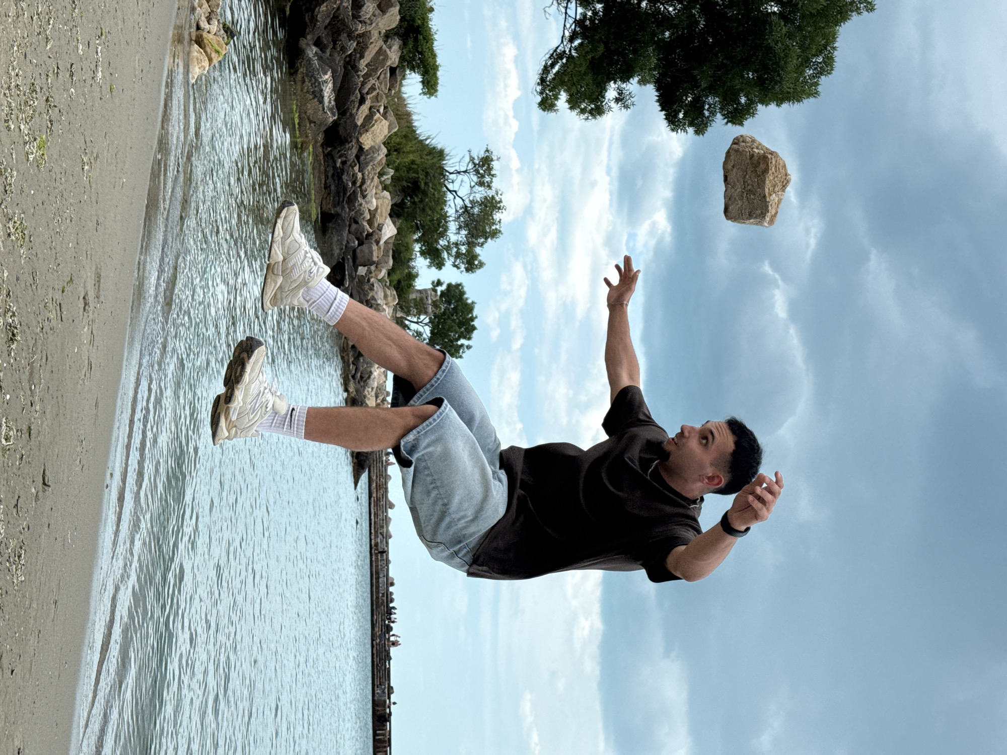 The image shows a man appearing to float in the air, seemingly defying gravity. He wears a dark t-shirt, light blue denim shorts, white socks, and white sneakers. His arms are outstretched, one hand reaching towards a levitating rock. The background reveals a coastal scene. To the left, there's a shoreline with water and rocks. The right side of the image displays a cloudy sky with a tree and more levitating rock. The image appears to be taken on the breakwater of the Varna Port, Bulgaria.