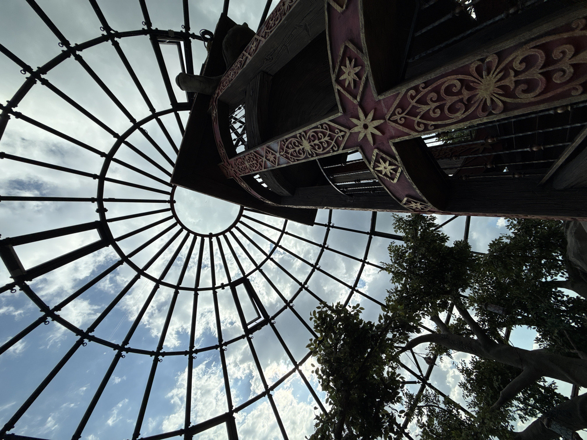 The image shows a low-angle view looking up at a geodesic dome structure made of metal beams and glass panels, through which a cloudy sky is visible. Inside the dome, a large, dark wooden tower with ornate red and gold decorative elements is visible. A spiral staircase can be seen within the tower. To the left of the tower, a large tree with green foliage is also visible, its branches reaching towards the dome. The setting appears to be an enclosed space, possibly an attraction or a themed structure. There are no people visible in the image. The location is specified as Simeiz settlement council, Ukraine.