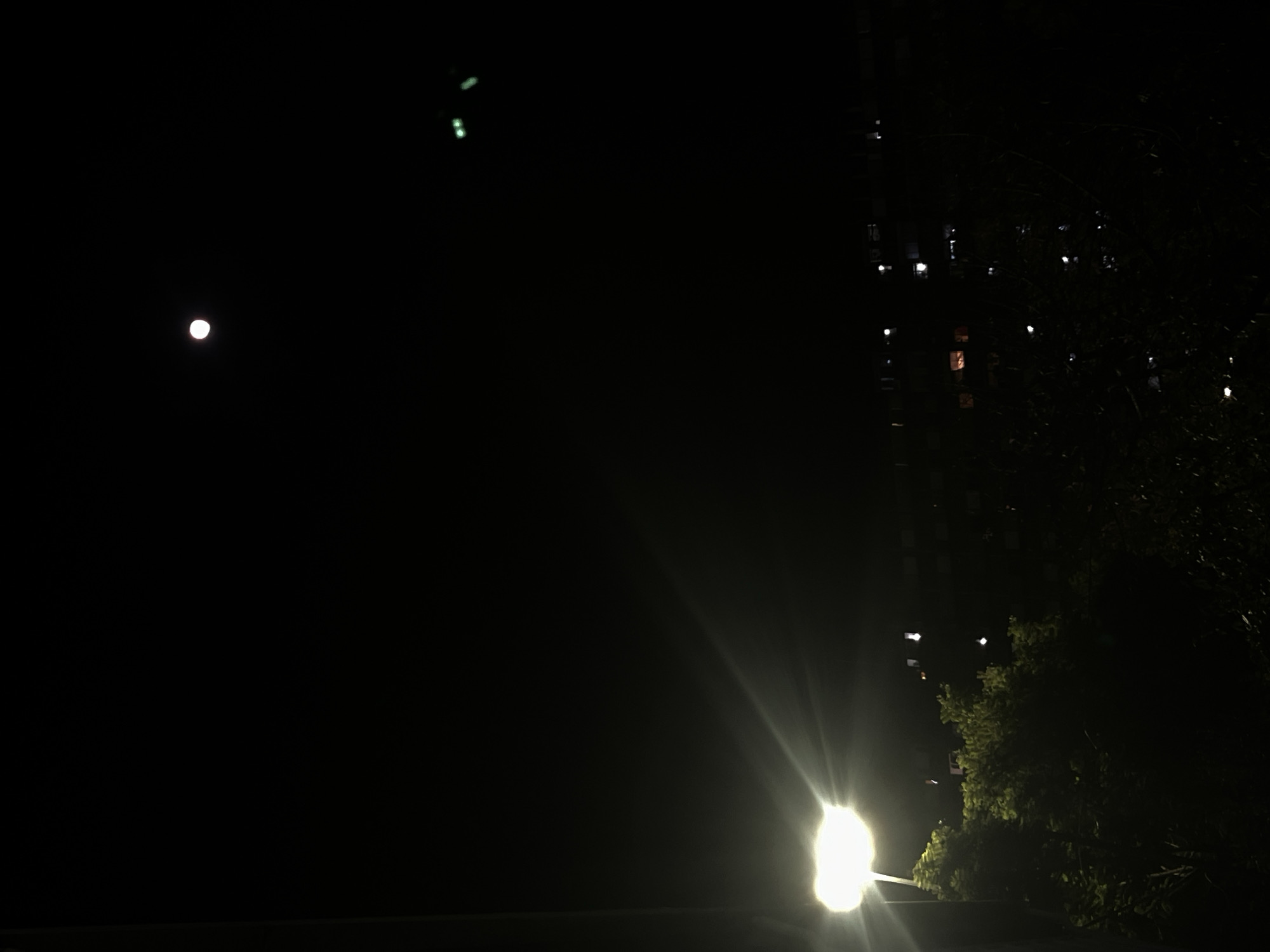 This nighttime photograph captures a serene urban or suburban landscape. The sky is dark, characteristic of evening hours, with a prominent, bright white Moon visible in the upper left quadrant. Higher up and slightly to the right, two small, faint green lights can be seen.
The lower right section of the image features a powerful artificial light source, likely a street lamp, which emits a significant lens flare, creating bright streaks radiating upwards and to the left. Adjacent to this light, the dark silhouettes of trees and foliage are visible, with some leaves subtly illuminated by the lamp's glow.
Further to the right, a multi-story building is partially visible in the darkness, distinguished by several illuminated windows, suggesting active occupancy. The overall scene depicts a quiet moment in a developed area, with natural elements blending with man-made structures and lights. No people or specific activities are discernible. The weather appears clear. There is no visible text in the image.