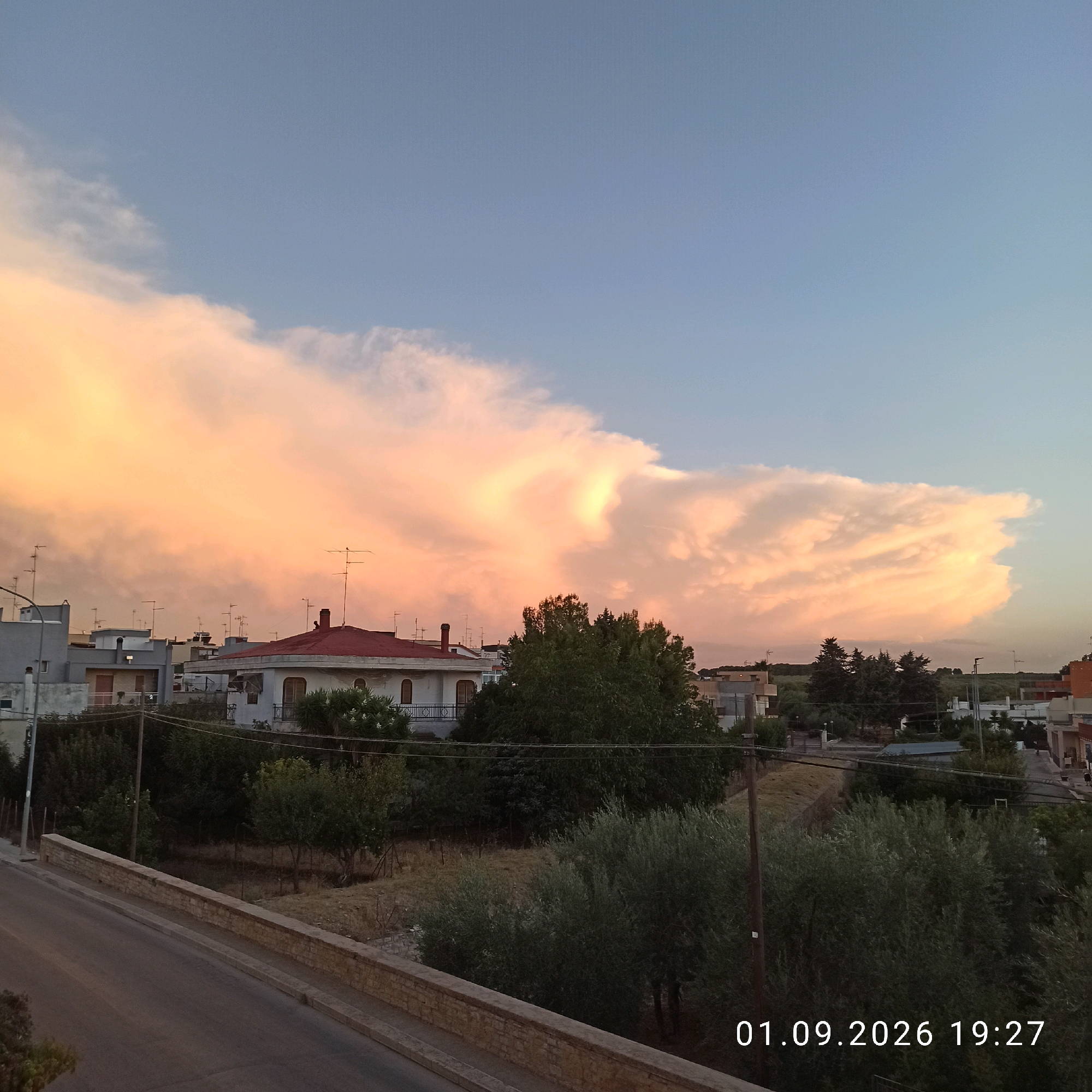 The image captures a dusky sky over the city of Bitonto, Italy, with large, billowy clouds illuminated by the setting sun, casting a warm orange and pink glow across the sky. The foreground features a road with a stone embankment and lush green trees and vegetation. Several buildings, including houses with red and white roofs, are visible in the mid-ground, along with numerous television antennas perched atop them. The overall scene is serene, suggesting a peaceful evening in a residential area. A date and time stamp, "01.09.2026 19:27", is present in the lower right corner of the image. There are no people or animals clearly visible in the frame.
