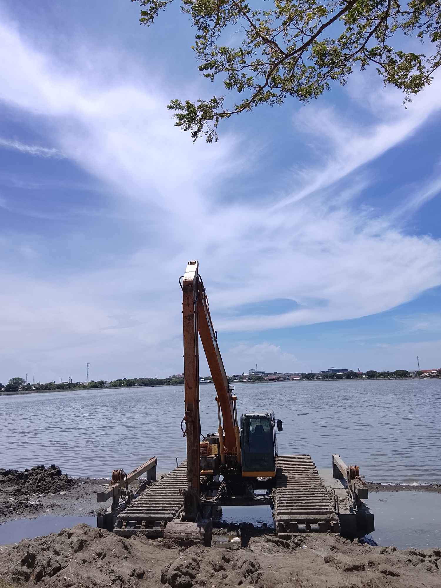 A yellow amphibious excavator is parked on a muddy shore beside a large body of water. The excavator has a long boom and a cab. The ground in front of the excavator is covered in mud and debris. The body of water appears to be a lake or a bay, with a distant cityscape visible on the opposite shore. The sky is blue with scattered white clouds. The scene is consistent with construction or land reclamation work in a coastal area.