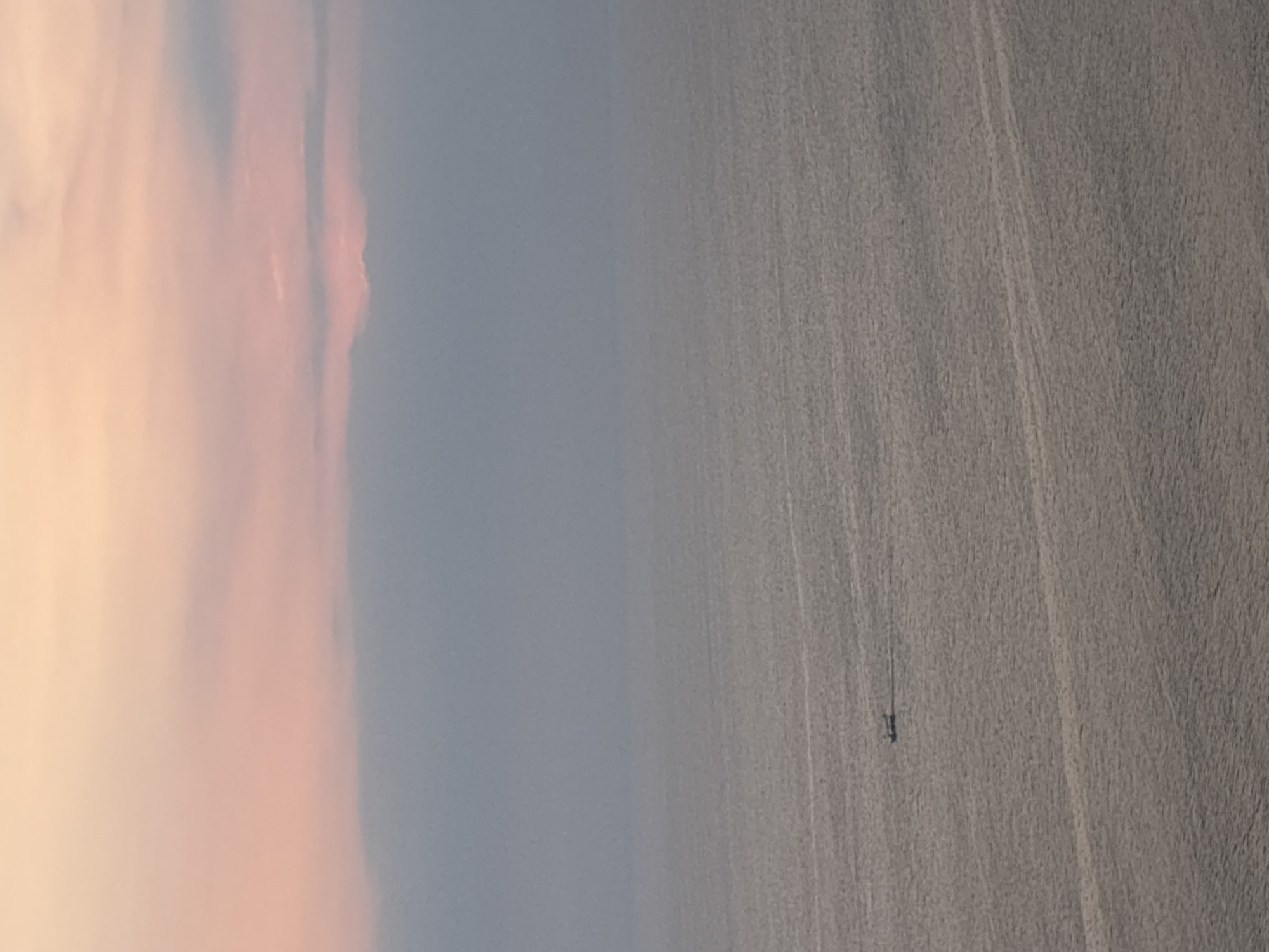 An aerial, wide shot captures a tranquil ocean scene under a soft, pastel sky at what appears to be sunset or sunrise. The sky transitions from a pale yellow at the top to soft pinks and then to a muted grey towards the horizon, with scattered, wispy clouds. The sea below is a calm, reflective grey, with subtle ripples and streaks of lighter color indicating gentle waves. A small, dark boat with a long wake is positioned in the lower left quadrant of the image, traveling from left to right. The overall impression is one of serenity and vastness. This scene is consistent with coastal views found in areas like Kuta Selatan, Indonesia, known for its beaches and ocean vistas.