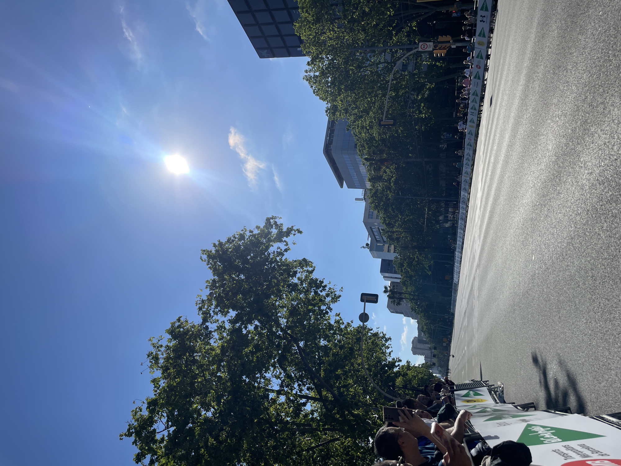 The image captures a scene in Barcelona, Spain, with a wide road flanked by trees and buildings. The sun shines brightly in a clear blue sky. A crowd of people lines the side of the road, many holding up their phones, suggesting they are filming or photographing an event. Barriers are set up along the roadside, with green and white signage visible. The road itself is wide and appears to be closed off for the event. Buildings of varying heights can be seen in the background, mixed with green trees. A traffic light is visible on the right side. The overall atmosphere is one of excitement and anticipation.