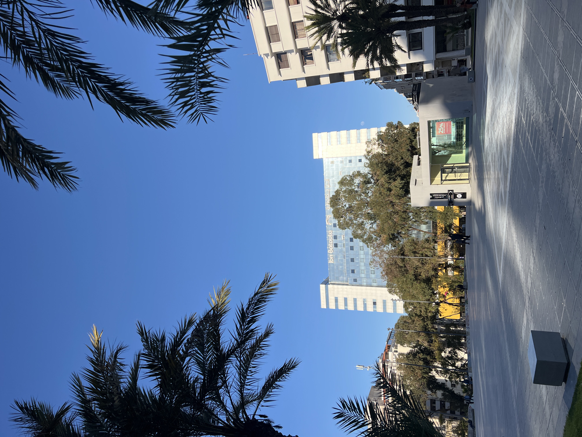 The image depicts a view in Tanger, Morocco. The frame is filled with palm trees in the foreground against a clear blue sky. Buildings of varying heights are visible in the background, including a taller, modern glass building with the logo "Ibn Battouta" and a light-colored building with multiple stories. The ground appears to be a patterned, paved surface suggesting a plaza or sidewalk. There are indications of trees lining the street. Shadows suggest a sunny day. There is some visible human presence, though details are obscured by distance and lighting.
