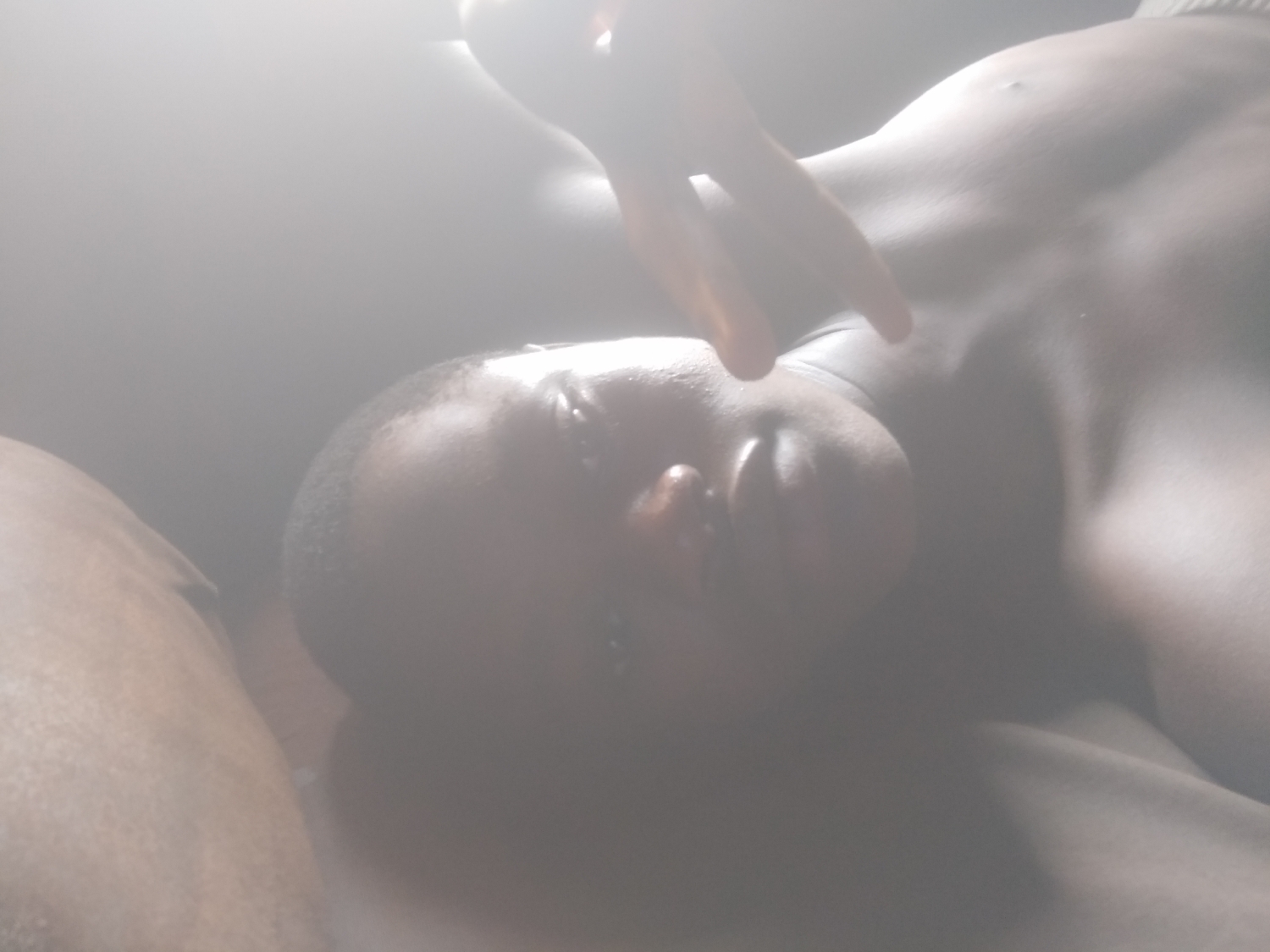 The image captures a person lying down, viewed from a slightly elevated perspective. The subject has dark skin, and their head is oriented towards the top of the frame, with the face looking upwards. A hand, likely belonging to the same individual, is visible near their head or face, partially obscuring the view. The upper torso of the person is also visible, suggesting a relaxed or resting posture.
The scene is dominated by an extremely bright, diffused light source, particularly concentrated in the upper and central parts of the image. This intense light creates a significant glare and a hazy, almost ethereal effect, making many details indistinct. The subject's facial features are somewhat obscured by this glare and the position of the hand. The strong lighting suggests either a bright daytime setting indoors, possibly near a window, or an area with very strong artificial illumination.
The background is largely indistinct due to the bright light and lack of focus, offering no specific cues about the location beyond an implied indoor setting. No specific objects are clearly discernible apart from the person's body parts. There is no visible text in the image. The overall impression is one of a serene, possibly contemplative moment, characterized by the striking and soft illumination.