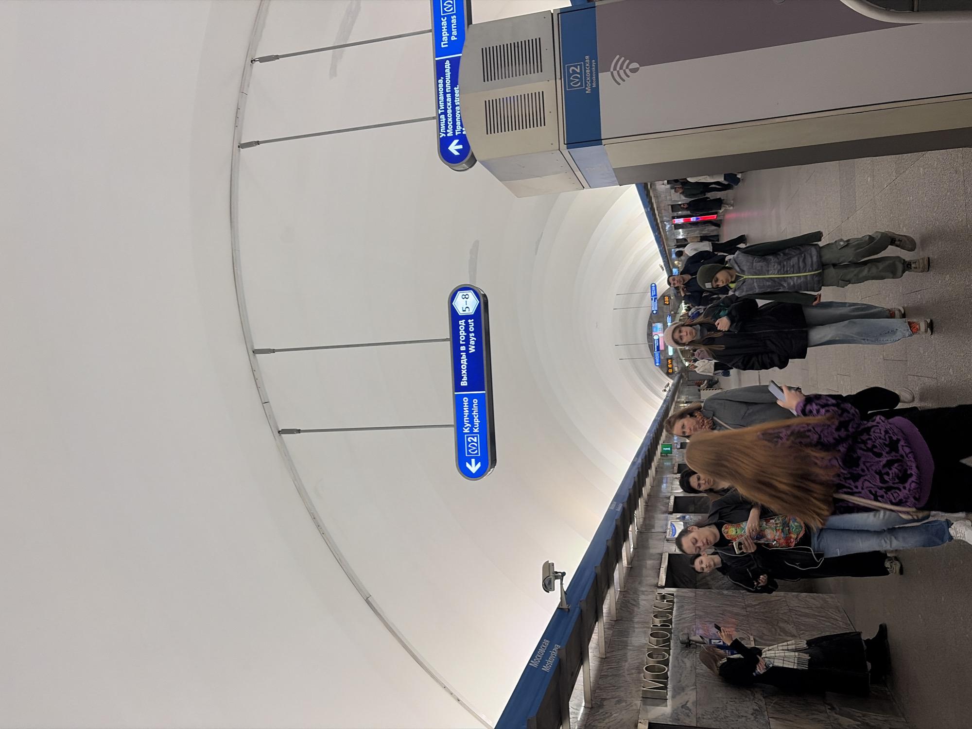 The image shows an underground metro station in Saint Petersburg, Russia. Passengers are visible on the platform, some looking at their phones. There are signs indicating the direction to Kupchino, Parnas, Ulitsa Tipanova, and Moskovskaya Square. The signs also show the line number of the metro (2). The name of the station, Moskovskaya, is visible on the wall. The architecture features a curved white ceiling with metal supports.