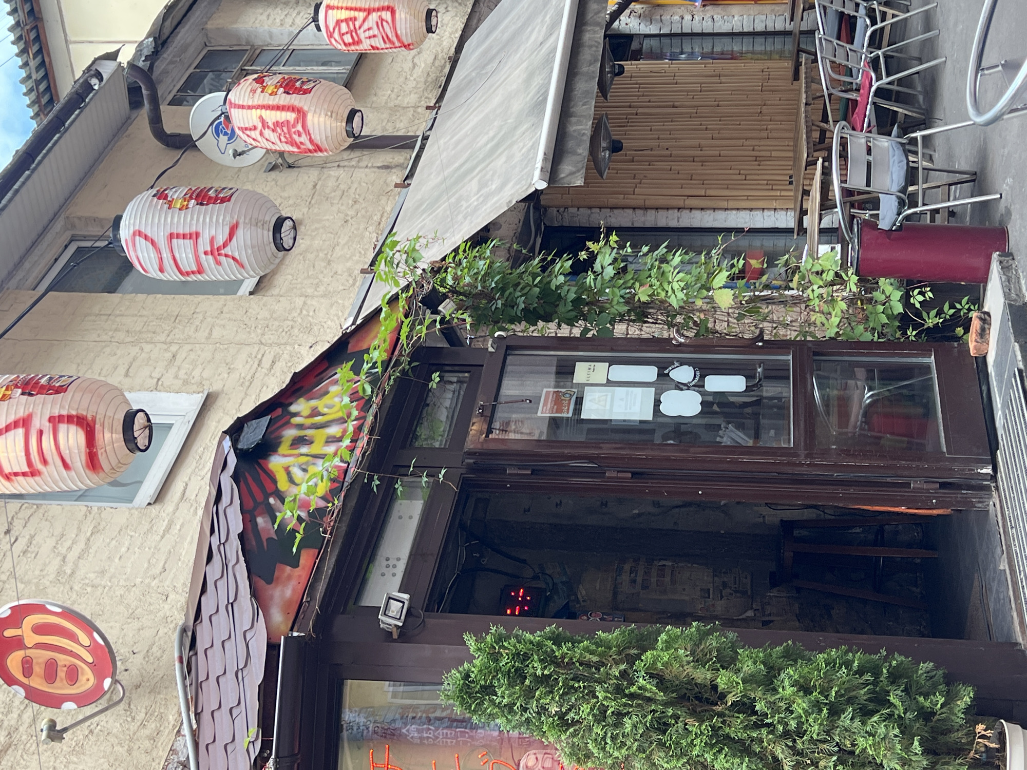 The image depicts the exterior of a Japanese restaurant located in Moscow, Russia. The restaurant's facade features a prominent dark brown wooden doorway with glass panels, through which the interior appears dimly lit, revealing a wooden stool and posters on the wall. To the left of the entrance, a tall, conical evergreen tree stands in a pot. Above the doorway, a brown tiled roof extends outwards, adorned with vibrant graffiti.
Several traditional Japanese lanterns, illuminated from within, hang from wires against the beige-colored building. These lanterns are cylindrical and white, decorated with red Japanese characters. One circular red sign with yellow markings is also visible on the upper left portion of the building.
The area in front of the restaurant is set up for outdoor dining, with several tables and chairs. The tables are made of wood, and the chairs are silver-toned metal with grey cushions. A grey awning extends over a section of the outdoor seating area. A tall, cylindrical red trash bin is situated near one of the tables. The building's exterior walls are a textured beige, with windows and what appears to be a bamboo-covered section on the right side. The overall atmosphere suggests a quaint, perhaps slightly weathered, establishment.