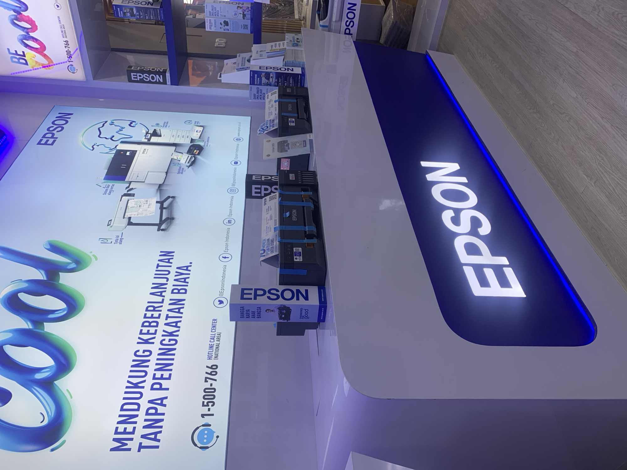 The image depicts an indoor retail or exhibition space, likely a store or a booth at a trade show, featuring products and branding associated with Epson. Prominently displayed on a large screen at the top is the word "COOL" in a stylized, bubbly font, alongside the Epson logo and the tagline "BE COOL". Below this, a large banner in Indonesian reads "MENDUKUNG KEBERLANJUTAN TANPA PENINGKATAN BIAYA," which translates to "SUPPORTING SUSTAINABILITY WITHOUT COST INCREASE."
A counter displays several Epson printers, some with packaging and marketing materials. The counter itself is white with blue accents and features a large illuminated "EPSON" logo at the bottom. To the right, on a shelf, are additional Epson products and promotional items. The overall color scheme is dominated by blue, white, and black, consistent with Epson's branding.
A prominent graphic on the large screen showcases various Epson printing technologies and a representation of the Earth, emphasizing global reach and sustainability. Contact information, including a hotline number (1-500-766) and social media handles like "@EpsonIndonesia" and "Epson Indonesia", are visible.
While the image does not explicitly state its location within Palembang, Indonesia, the presence of the Indonesian language and local contact numbers strongly suggests it is a retail or promotional space within the country. The environment appears to be clean, modern, and well-lit, typical of a commercial establishment. There are no people clearly visible interacting with the products, suggesting the photo was taken during a quieter period or that the focus is on the display itself.