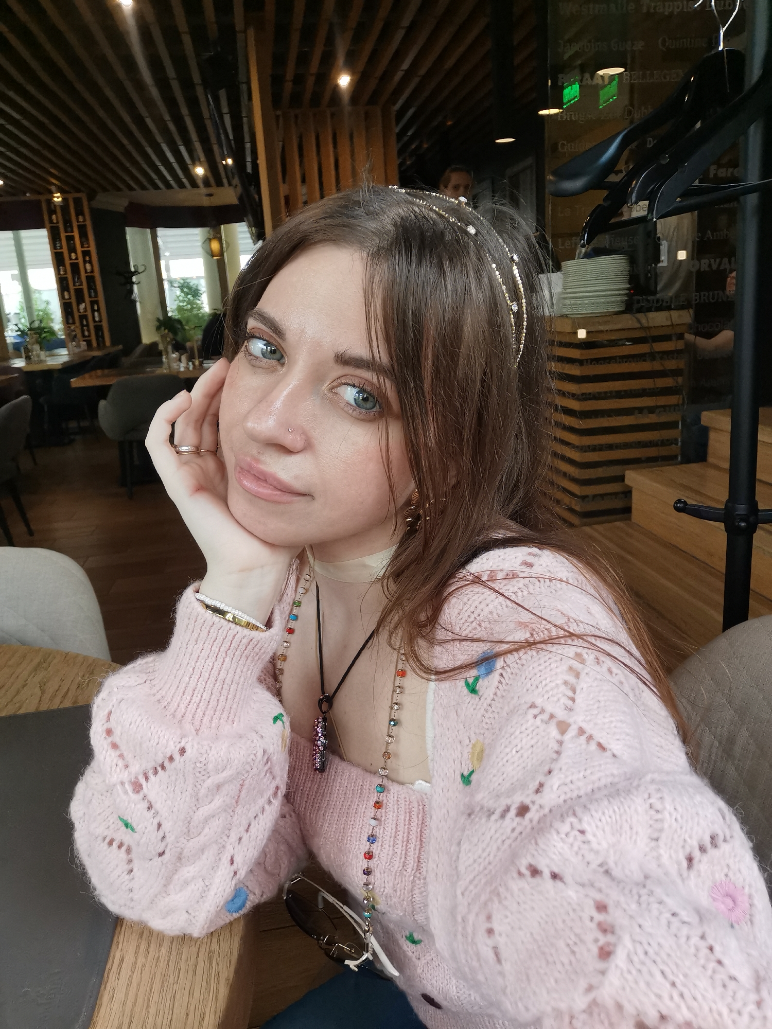 The image shows a young woman indoors, presumably in a restaurant or cafe. She has long brown hair, blue eyes, fair skin, and is wearing a jeweled headband. She is wearing a light pink sweater with small floral embroidery. Her left hand is resting on her cheek, and she is looking directly at the camera.
Several necklaces are visible around her neck, along with a choker. A ring is on her finger, and a bracelet on her wrist.
The background shows a wooden ceiling, shelving with bottles, tables, and chairs, suggesting a dining area. Outside the window is a view of greenery. A menu board with writing in French and English is partially visible. This menu features multiple belgian beers.